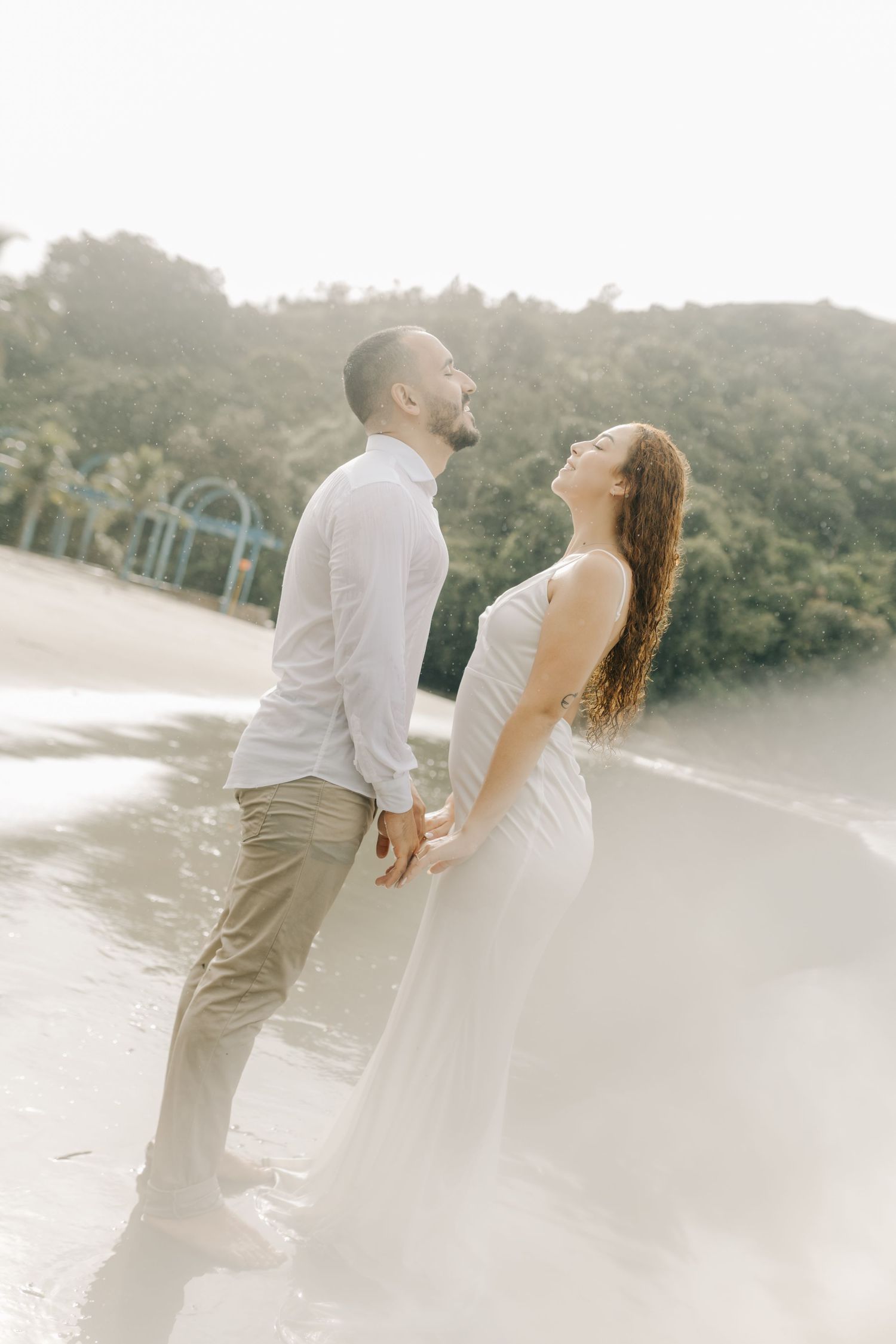 Romantic couple in white attire shares intimate moments on a misty beach at sunset.