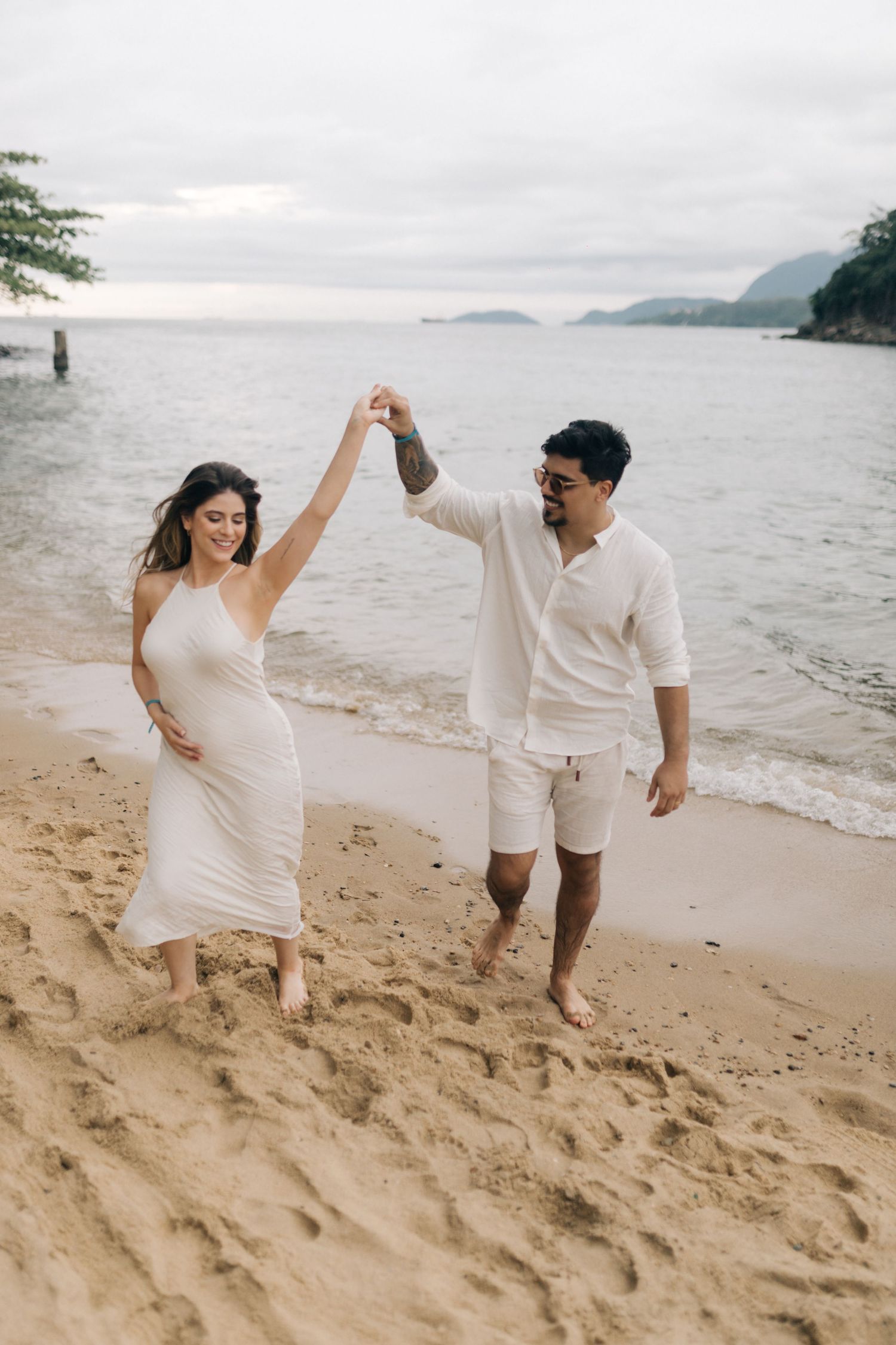 A couple in matching white outfits dancing together on a beach with mountains in the distance.