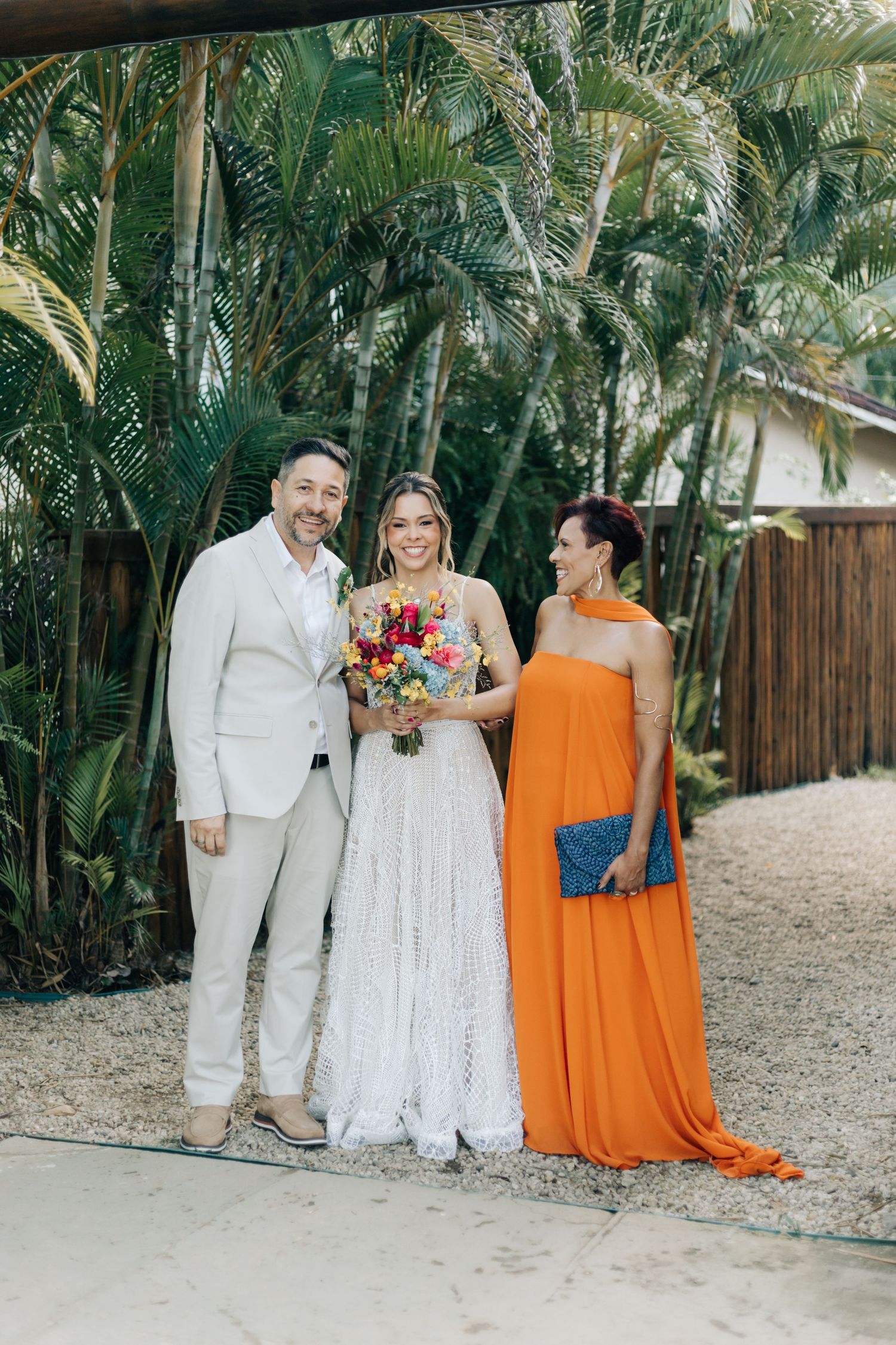 Wedding group photo with coordinated white and vibrant orange outfits in a tropical garden setting.