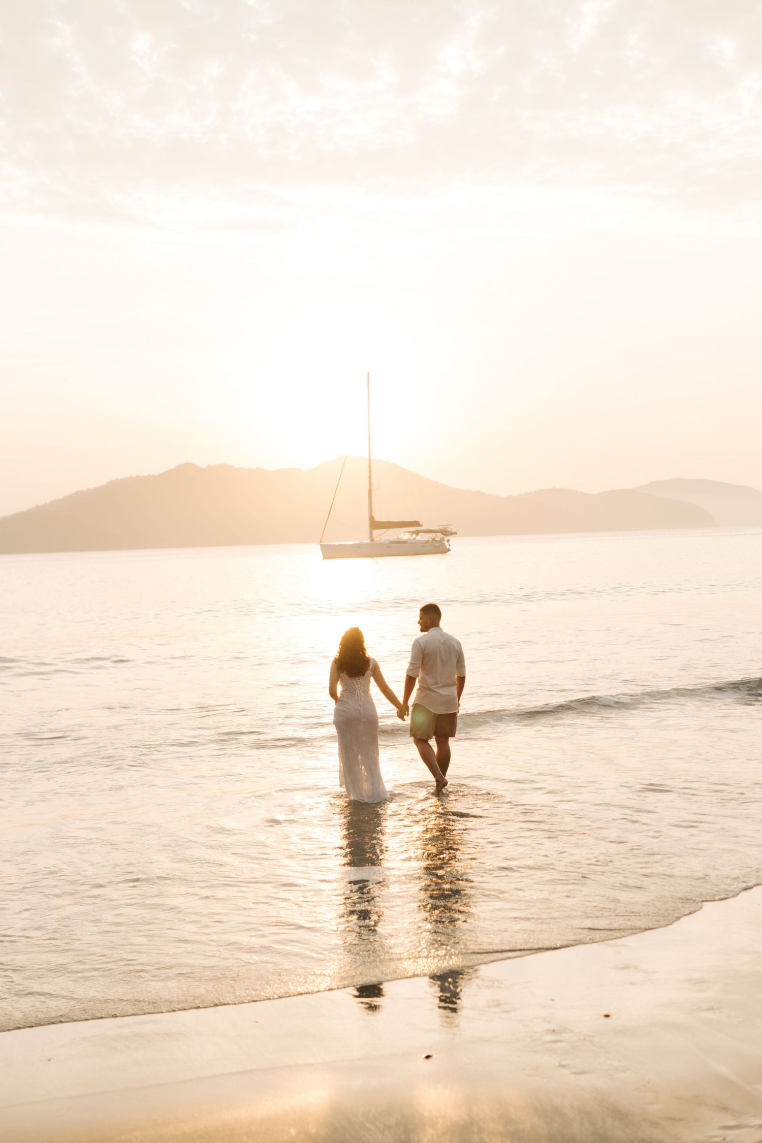 A couple walks hand in hand in shallow ocean water with a sailboat on the horizon at sunset.