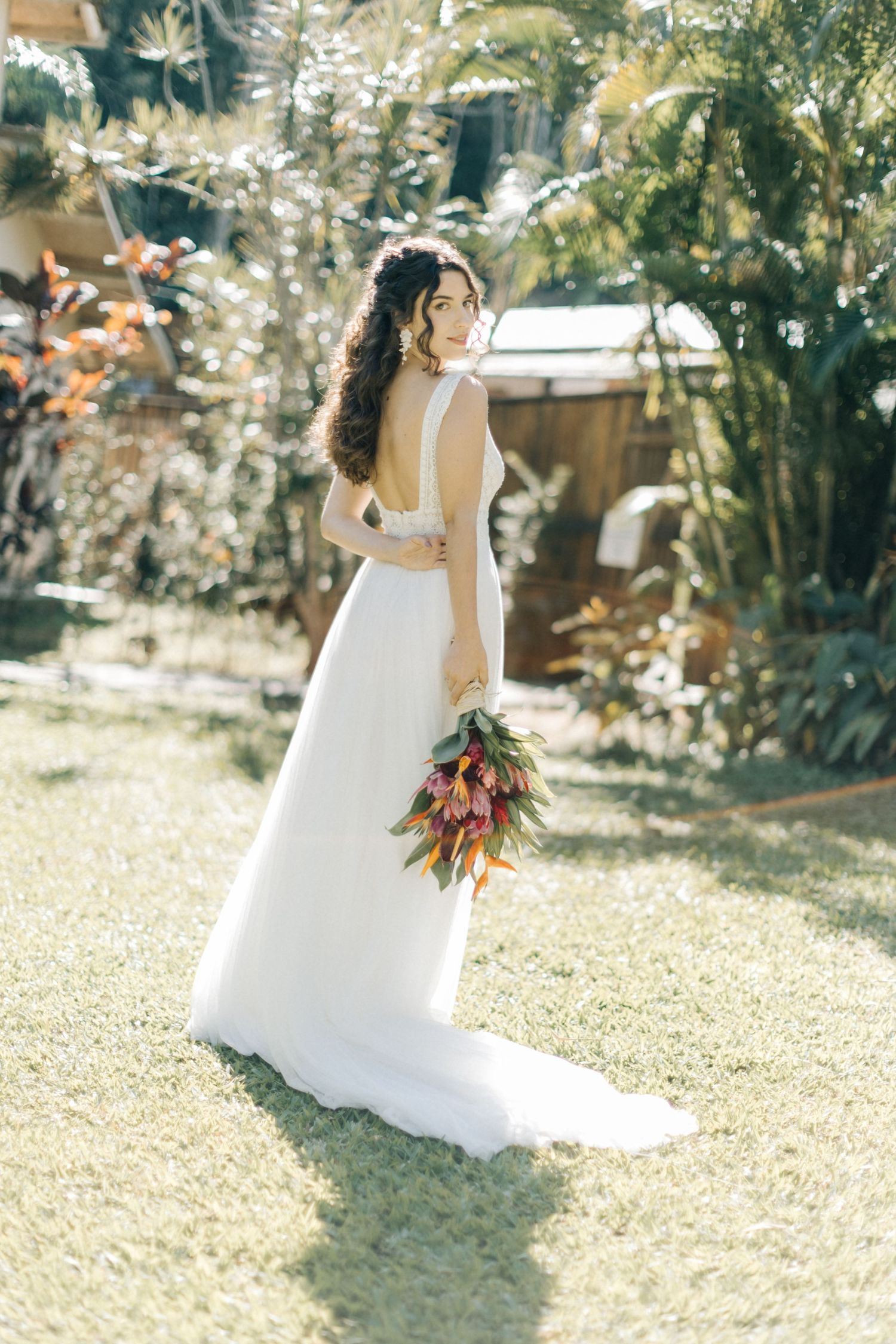 Back view of a bride in a flowing white gown holding a colorful bouquet in a sunlit garden.