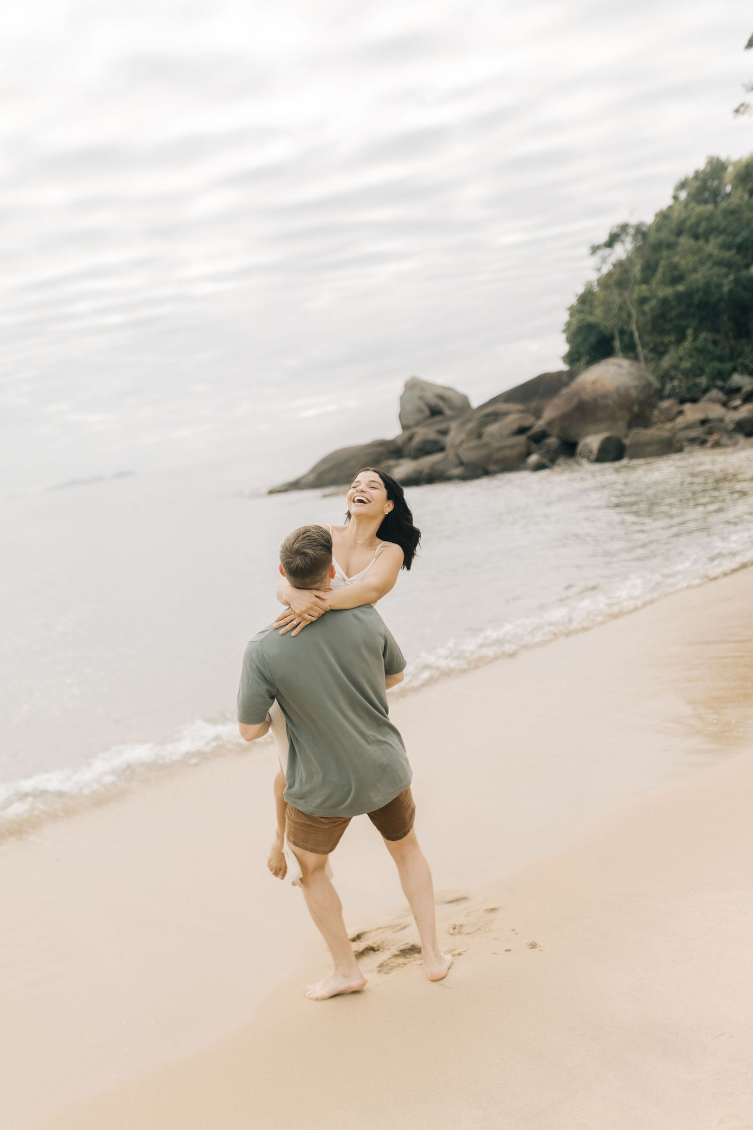 A couple embraces playfully on a scenic beach with rocky outcrops and overcast skies.