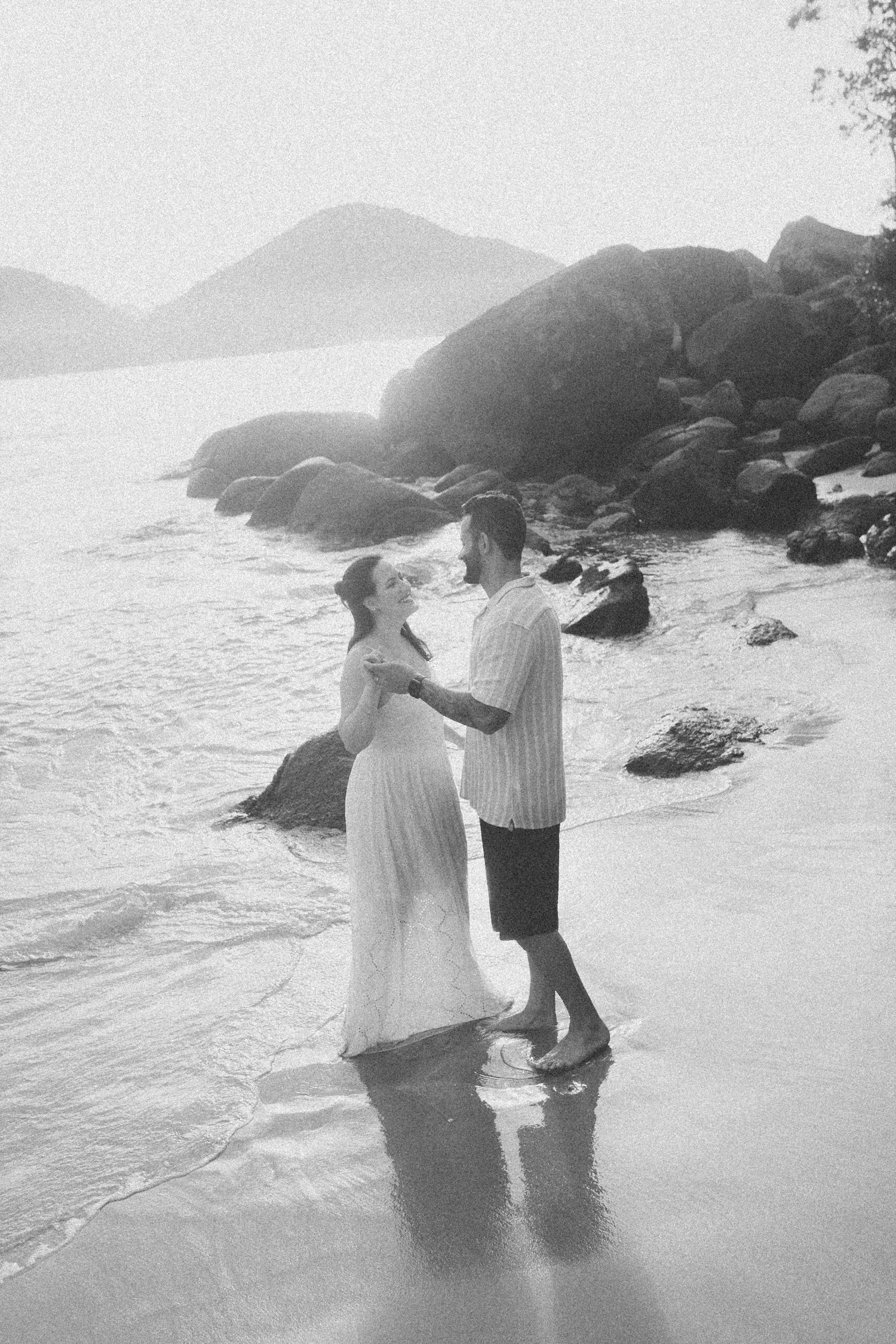 Black and white beach portrait of couple standing in gentle surf near rocky shore.