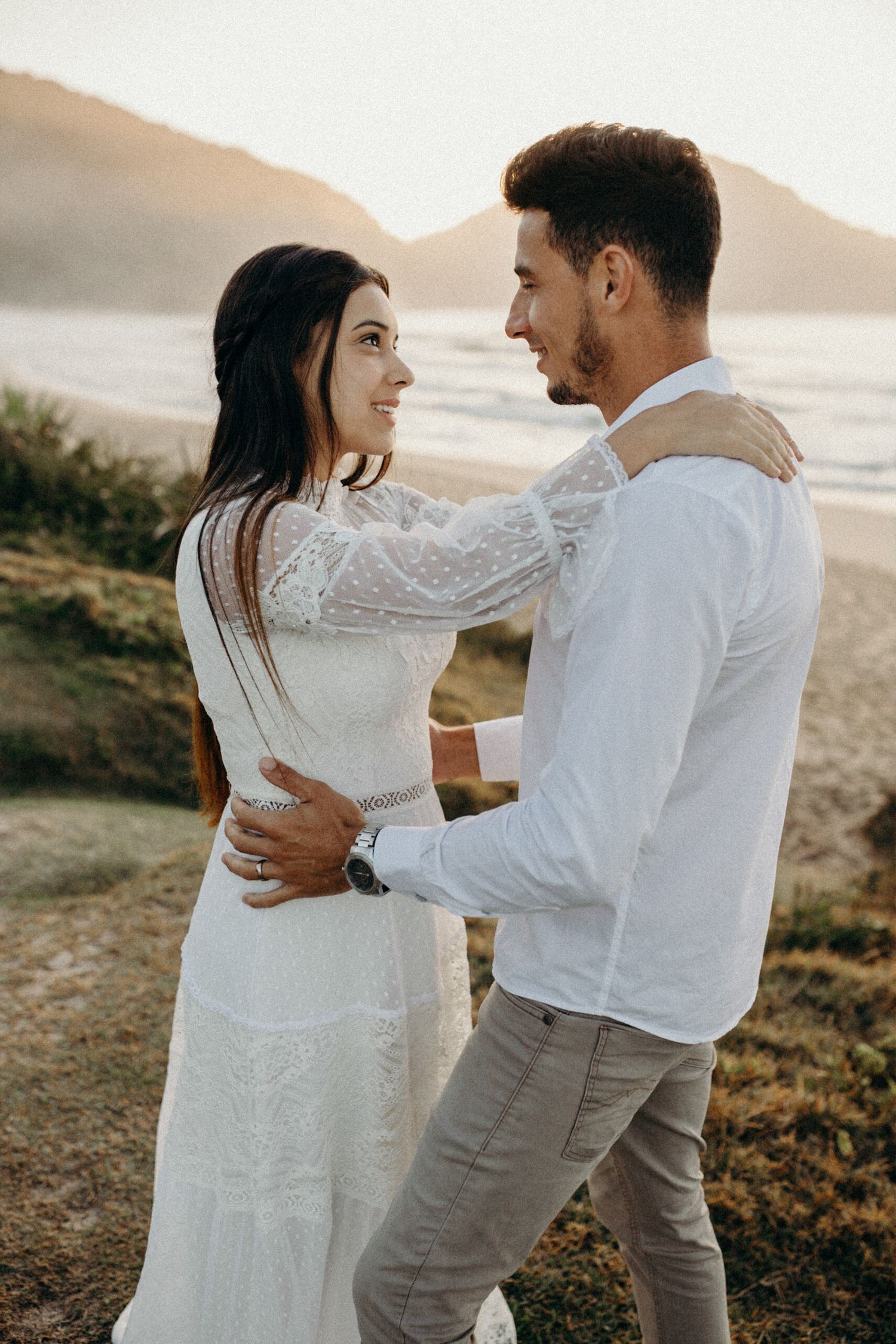 Romantic couple in white embraces on a beach during golden hour.
