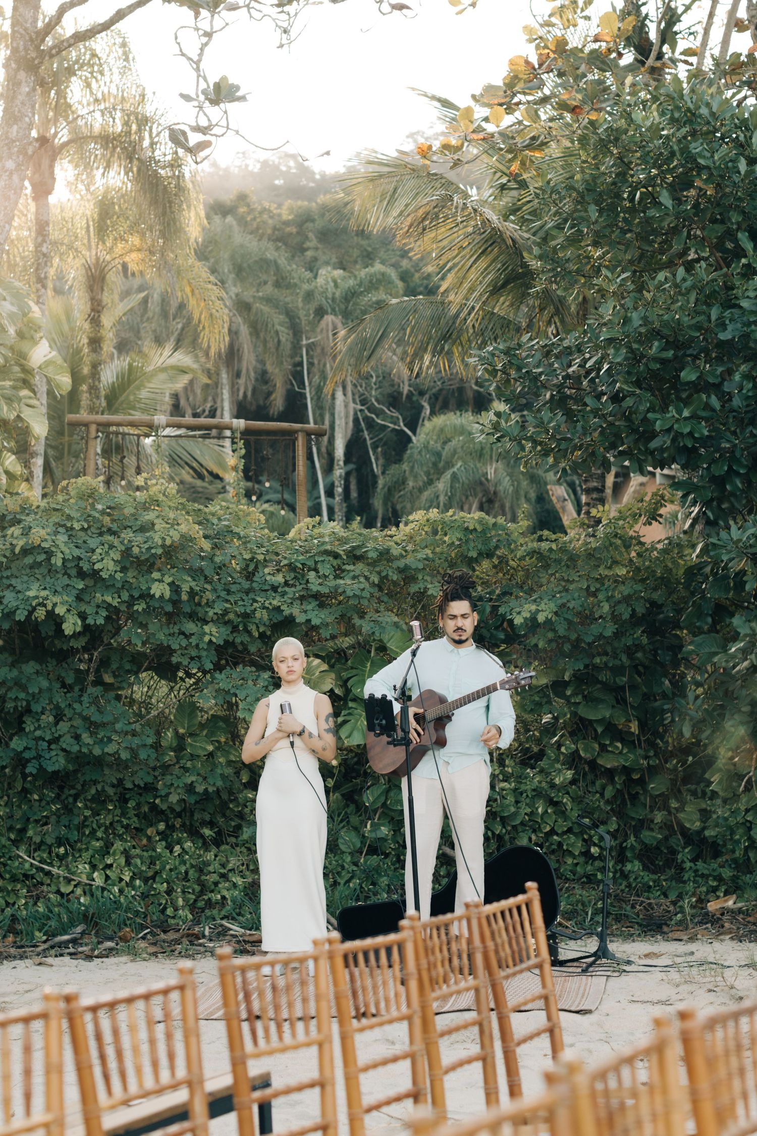 Musicians perform at an outdoor wedding venue surrounded by tropical palm trees and wooden chairs.