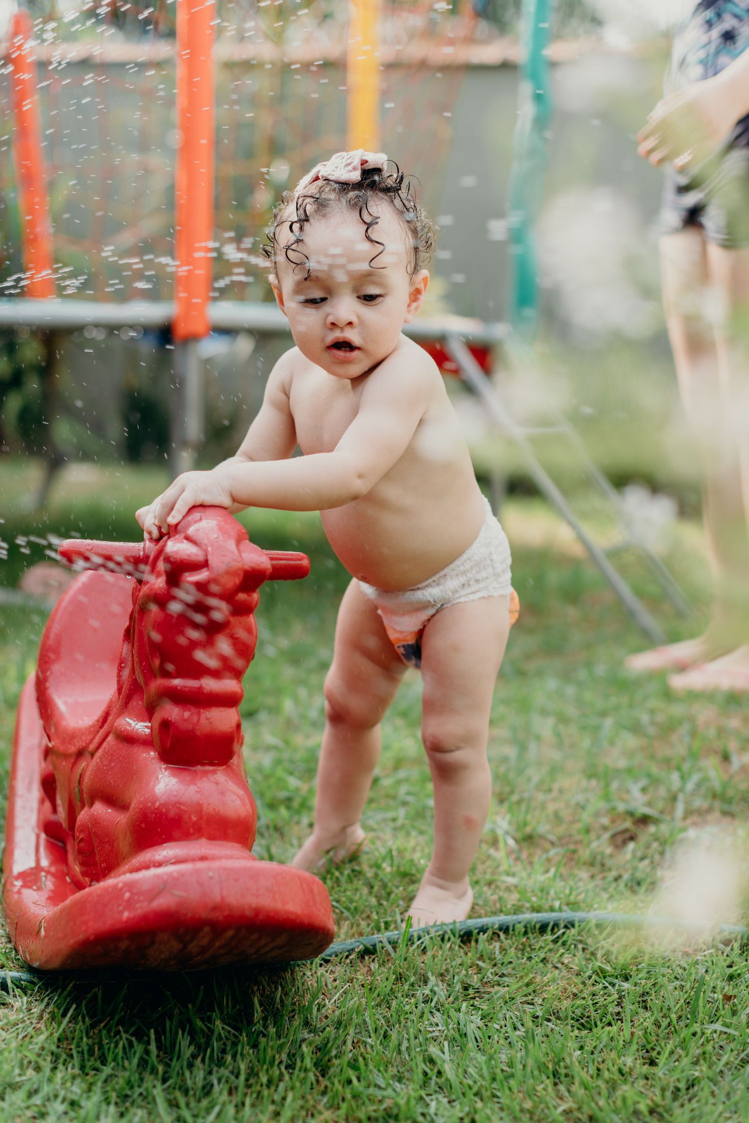 Small child in a diaper exploring a red plastic watering can on grass.