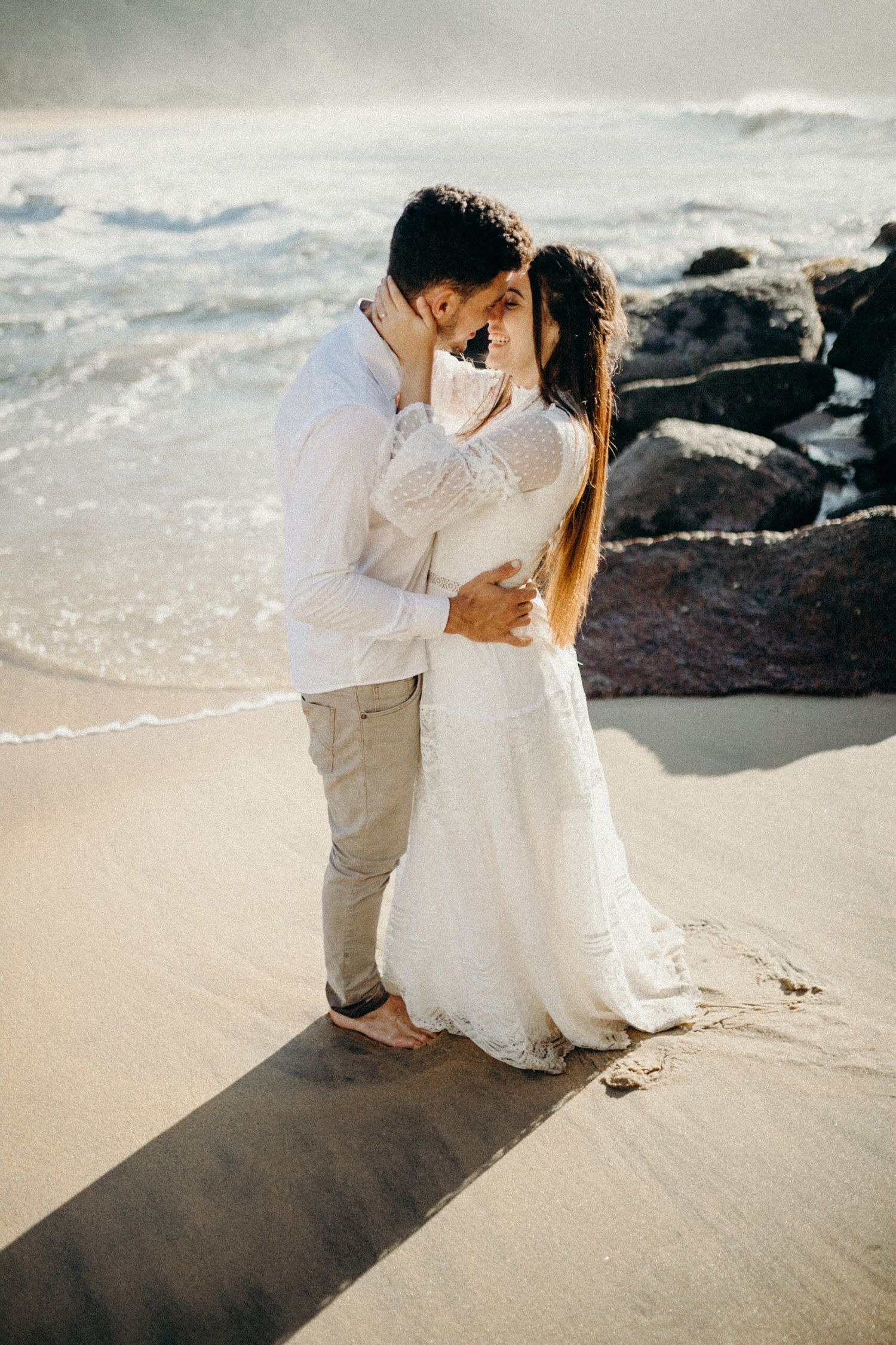 A couple embraces on a sandy beach with crashing waves and dark rocks creating a dramatic backdrop.