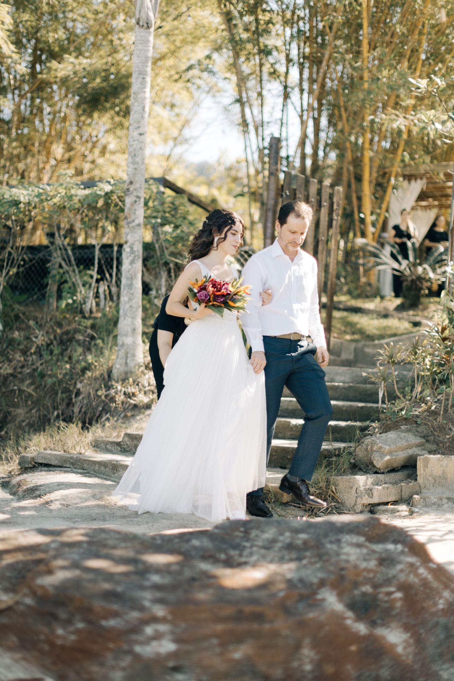 A couple walks along a stone path surrounded by trees at an outdoor wedding venue.