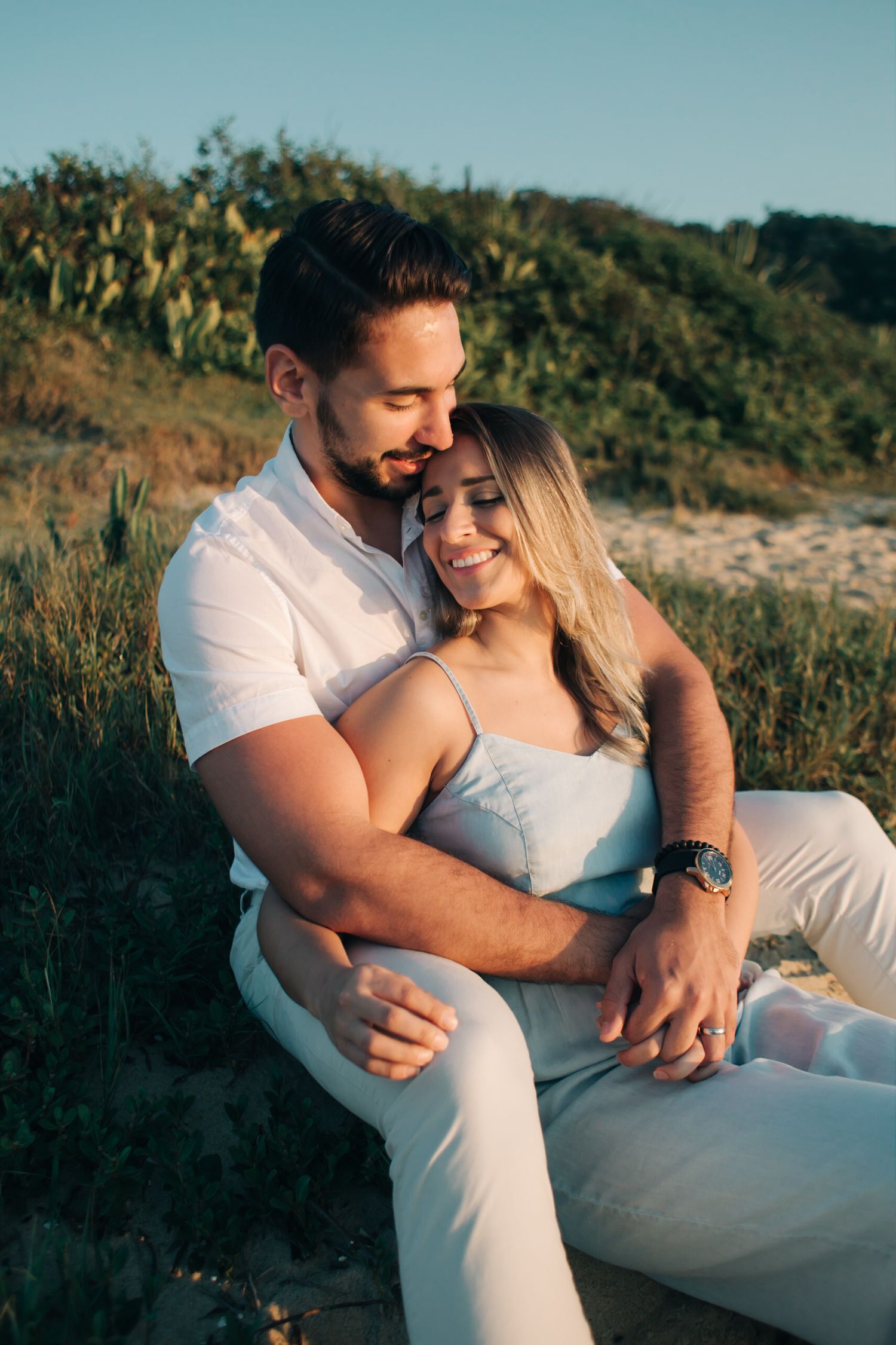 Couple in coordinated white outfits embracing while sitting outdoors against a natural backdrop.