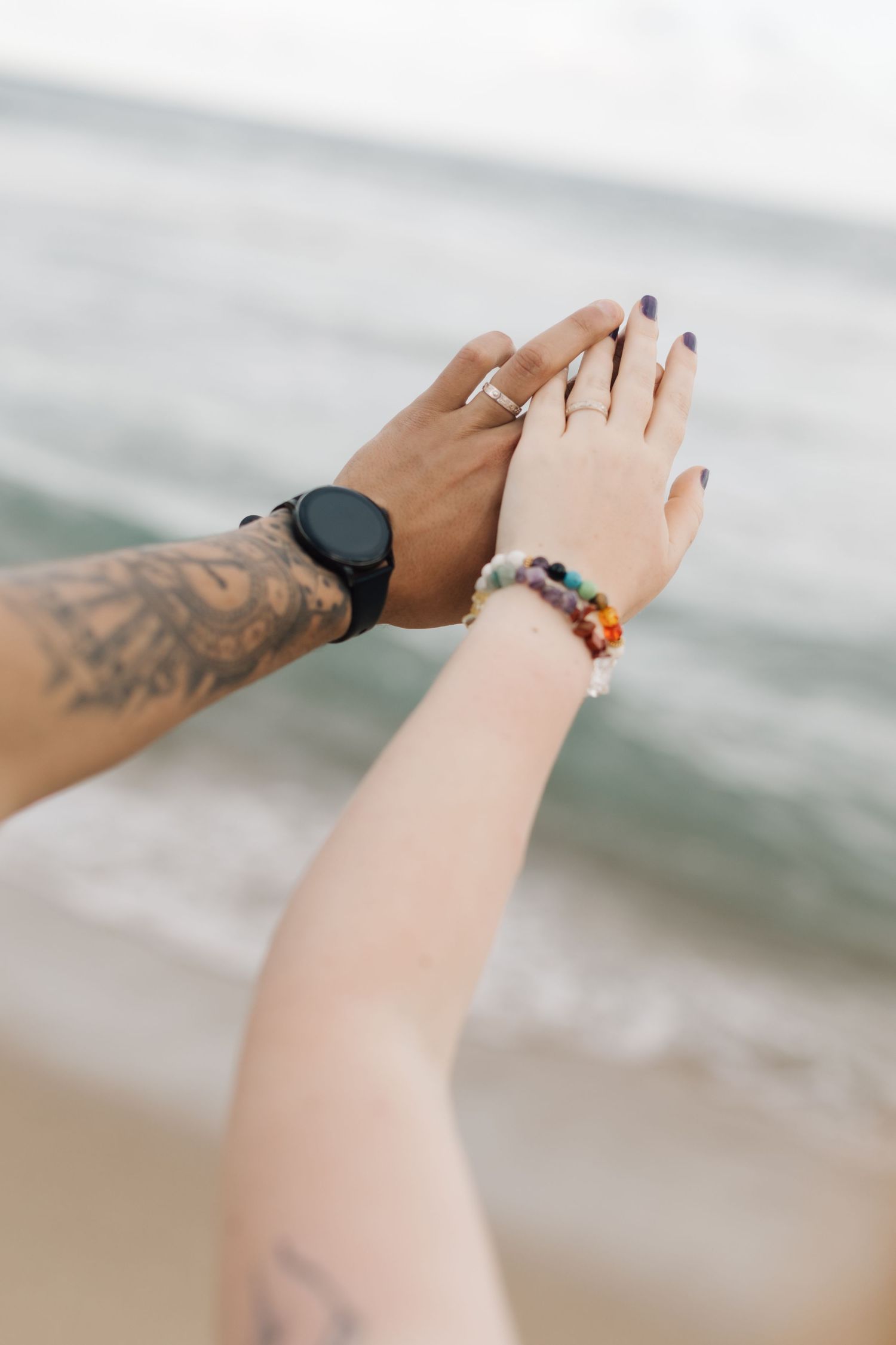Close-up of hands with tattoos and jewelry against an ocean background.