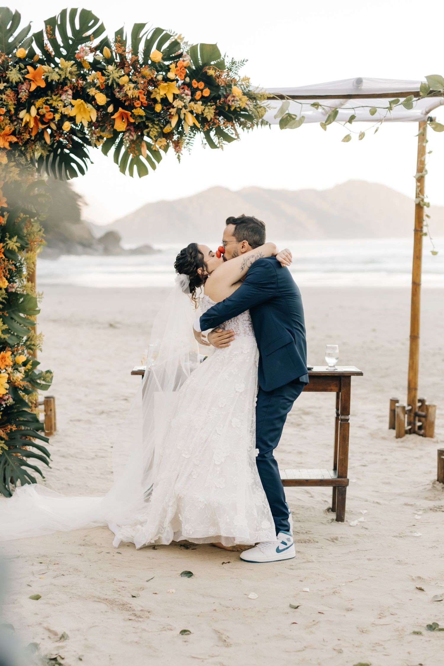 Romantic beach wedding embrace under an arch decorated with orange flowers and monstera leaves.