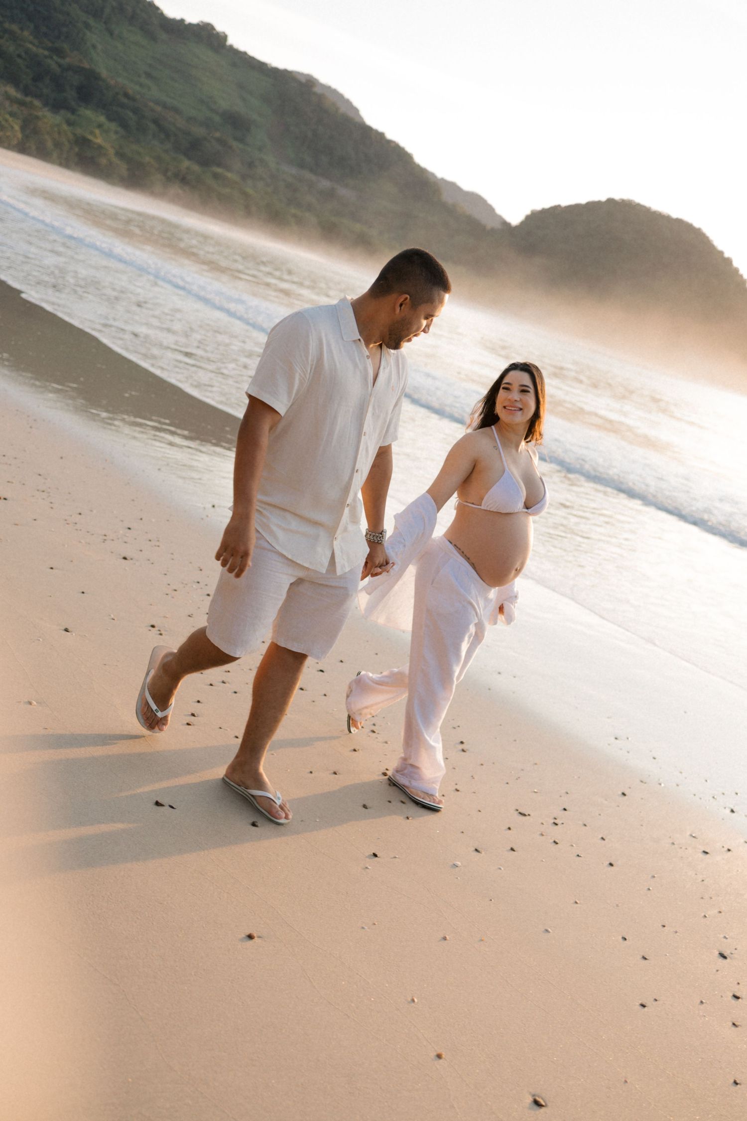 A couple runs playfully along the shoreline at sunset wearing matching white outfits.