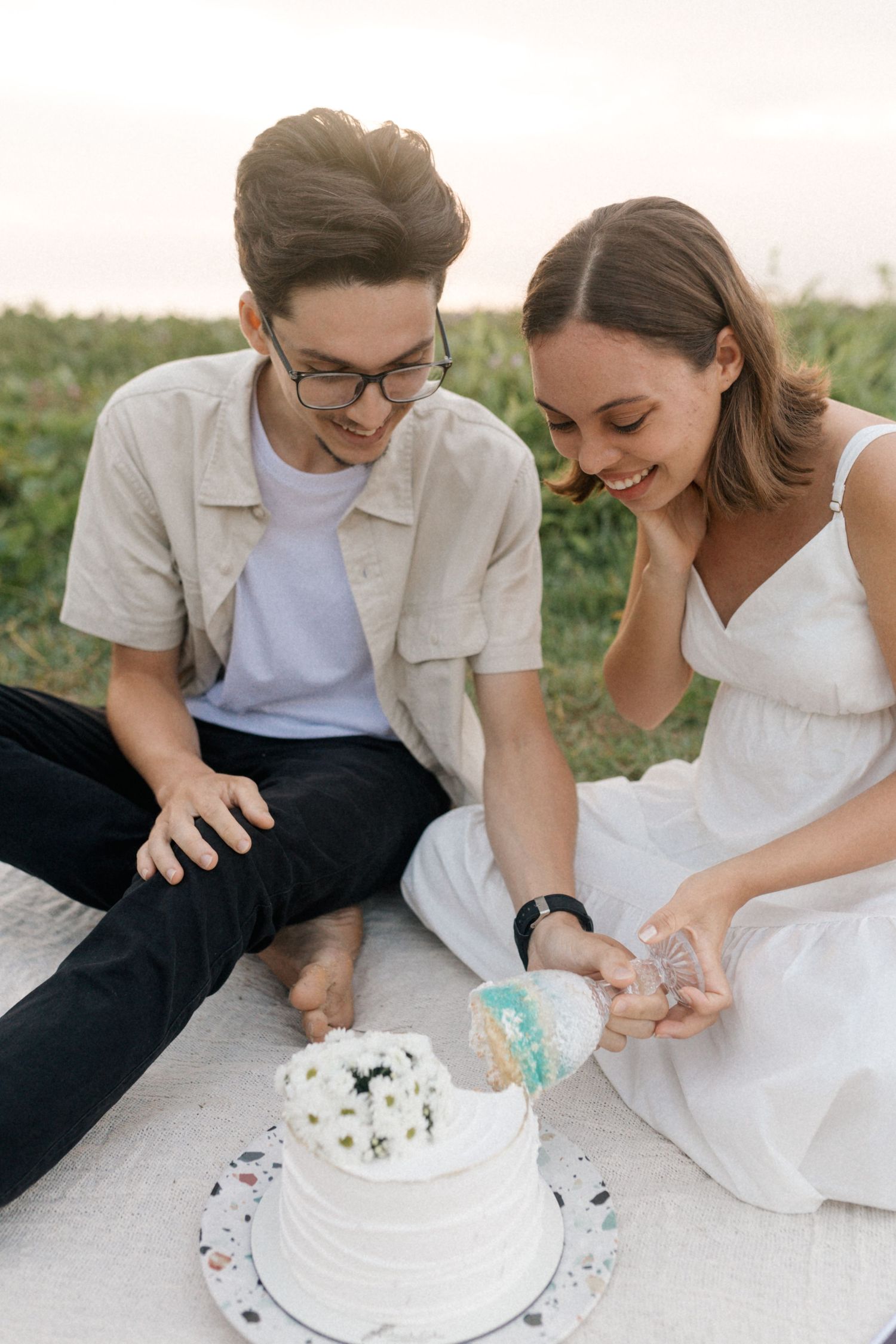 Couple playfully decorating a white cake while sitting on picnic blanket outdoors.