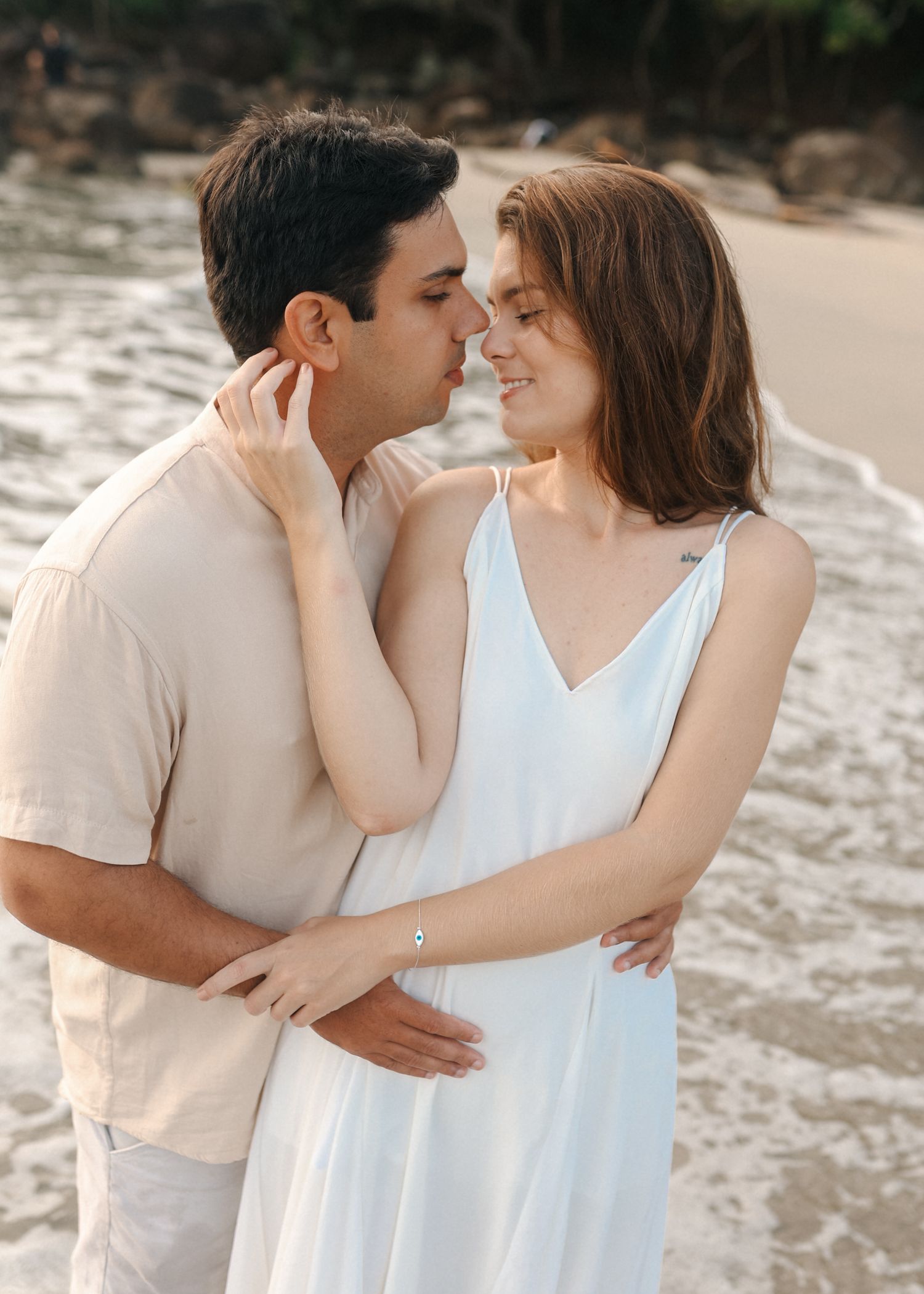 An intimate moment shared between a couple in light colored clothing on a beach at sunset.