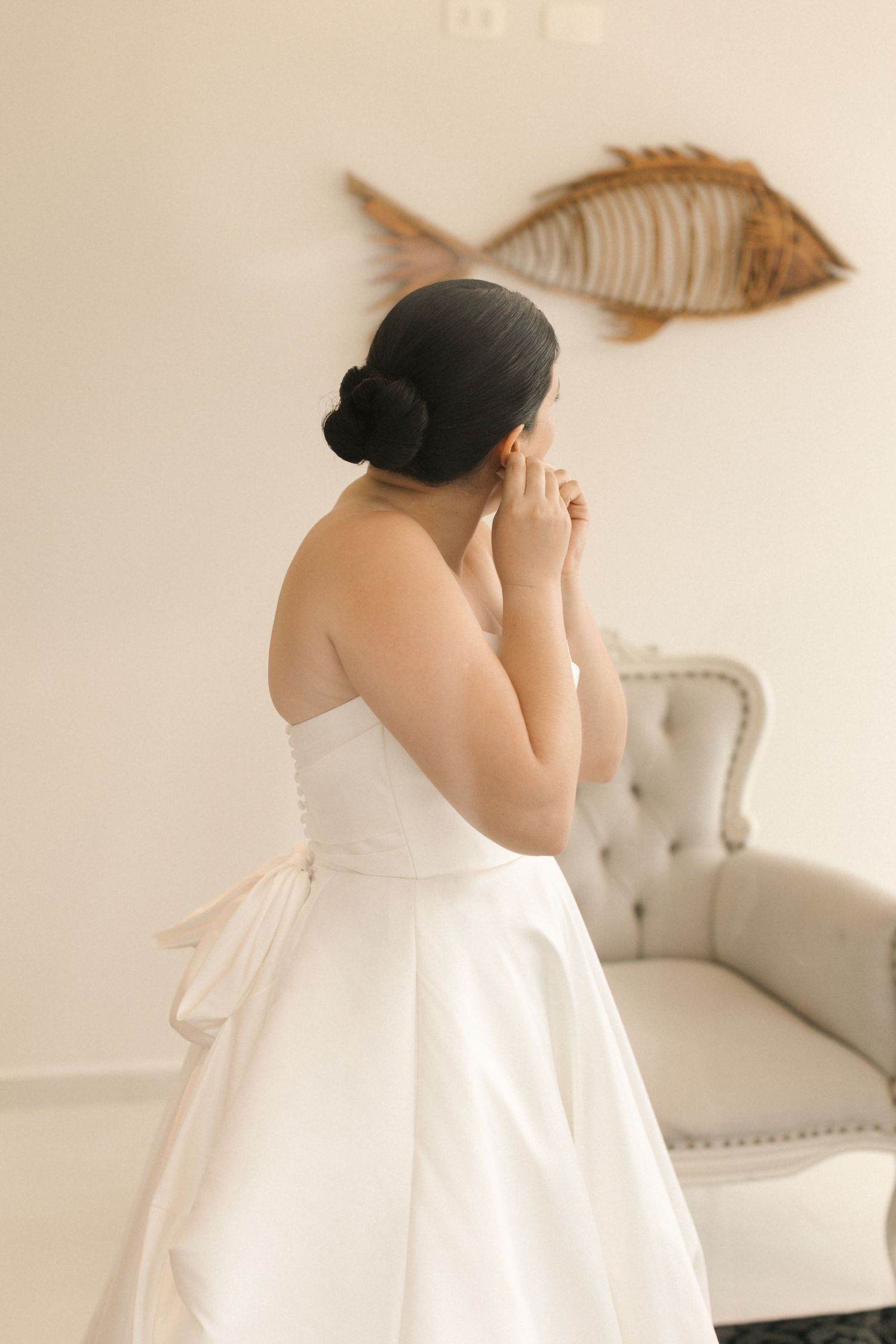 Back view of bride in white wedding gown standing near vintage chair with fish artwork on wall.