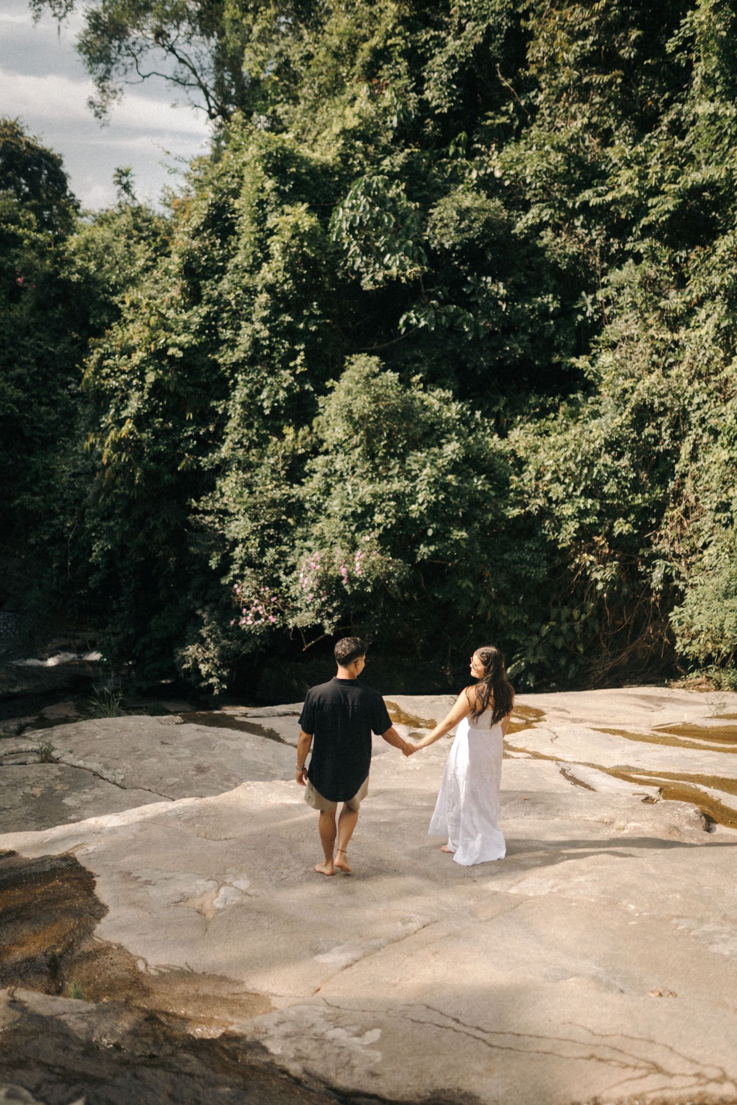 Couple walking hand in hand on a rocky outcrop surrounded by lush forest.