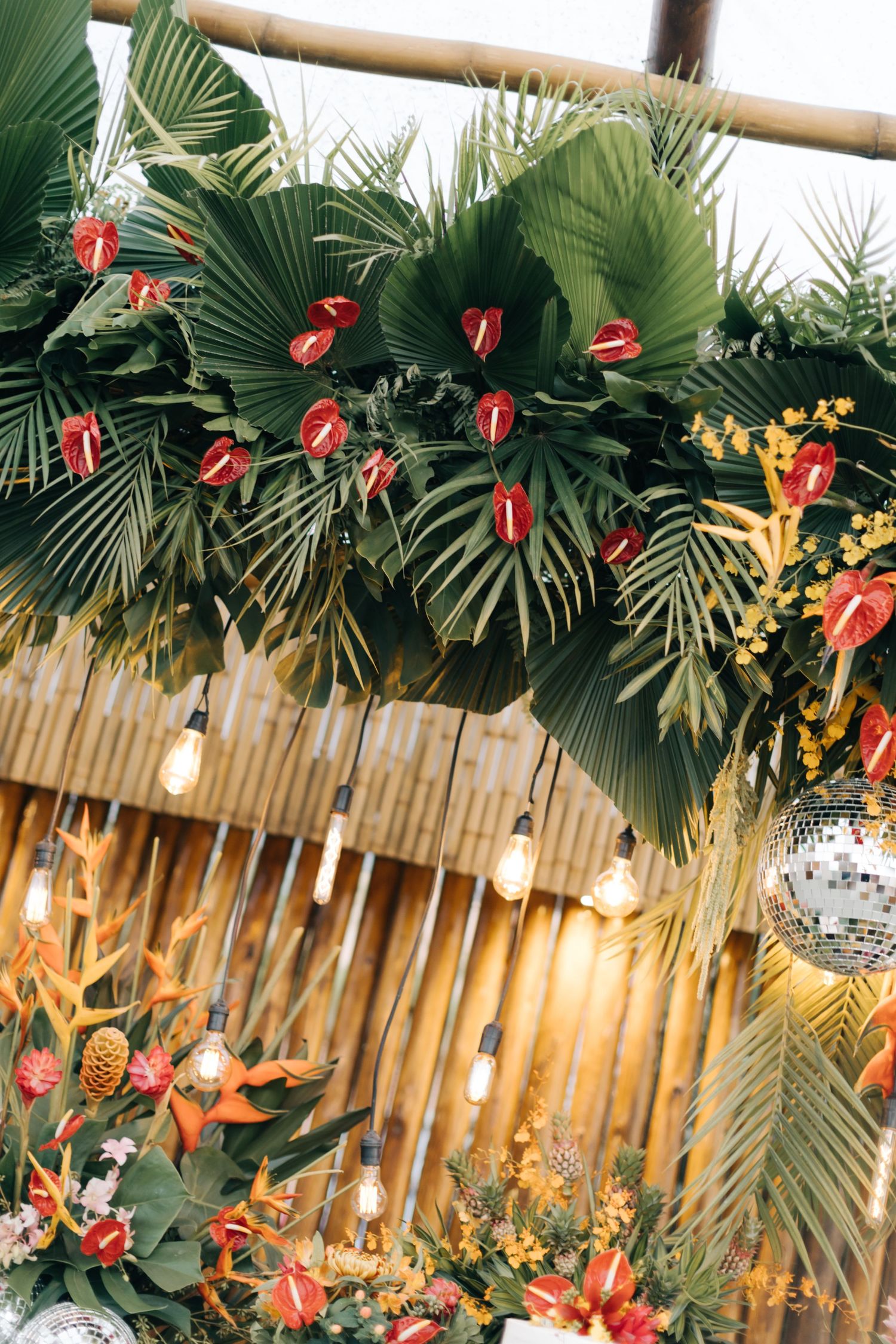 Palm fronds and red anthurium flowers decorating bamboo ceiling with hanging Edison bulbs.