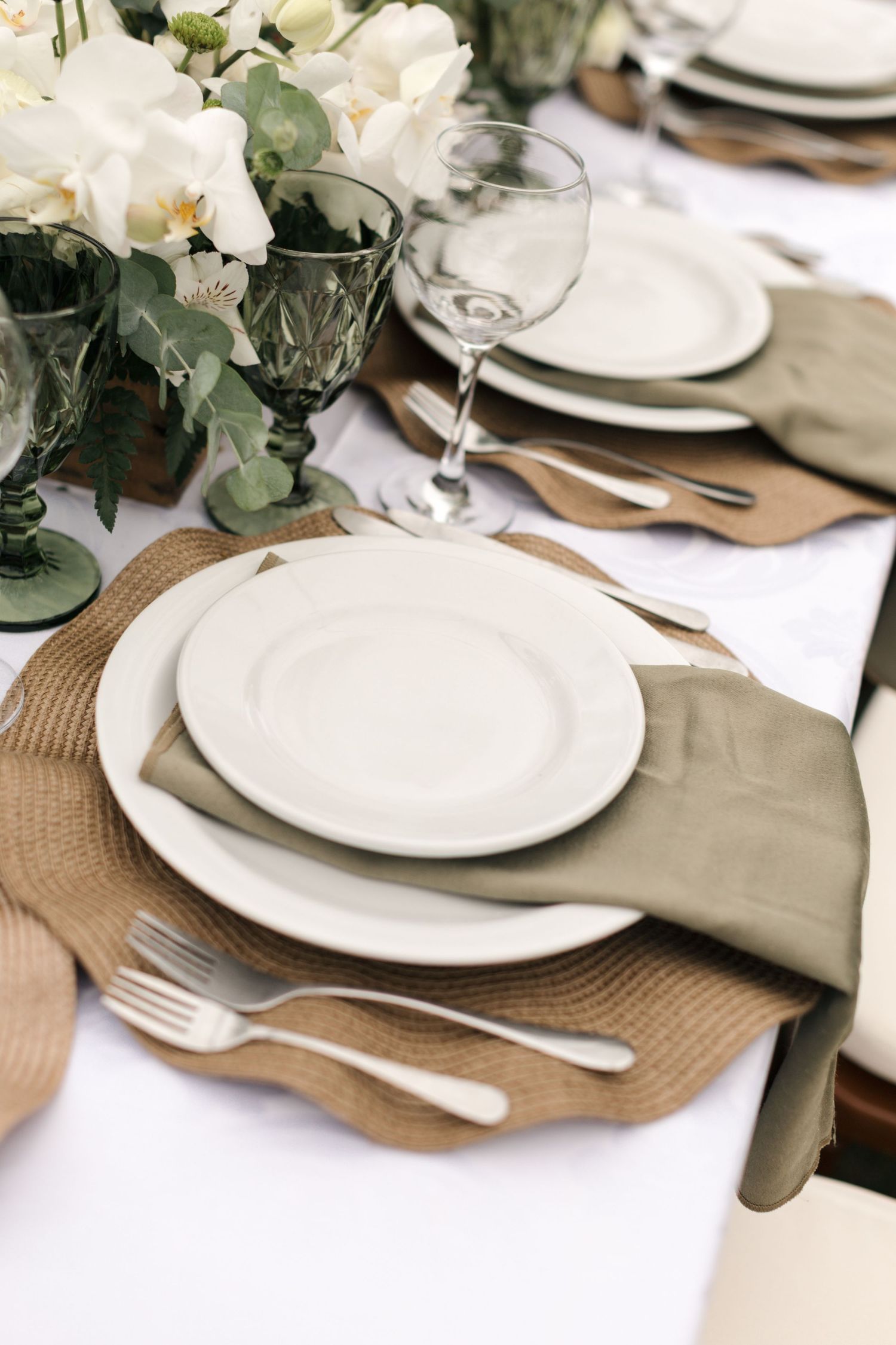 Elegant table setting with white plates, sage napkins, and woven placemats on a crisp white tablecloth.