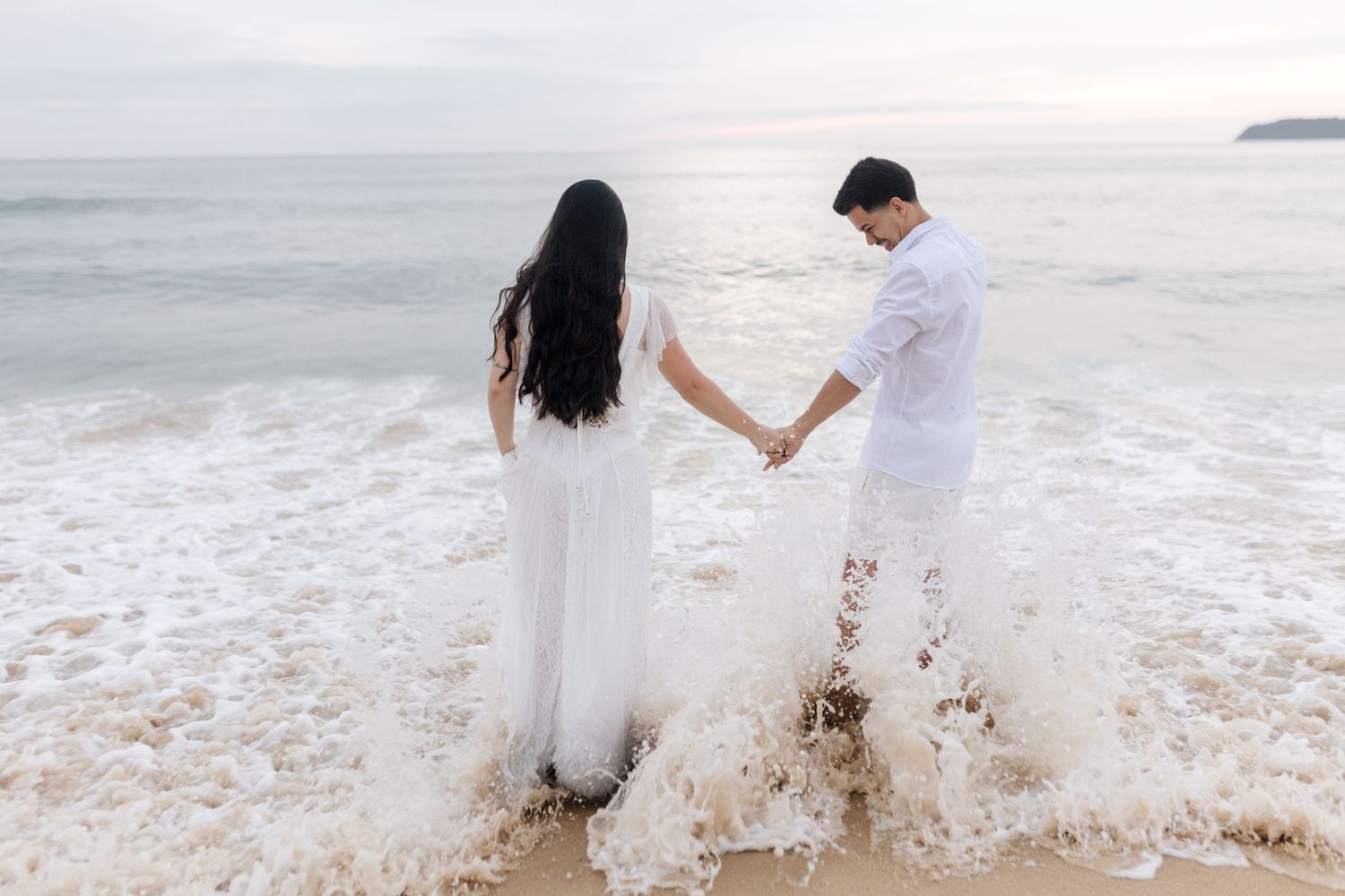 Romantic couple in white outfits walking and embracing on a beach at sunset.