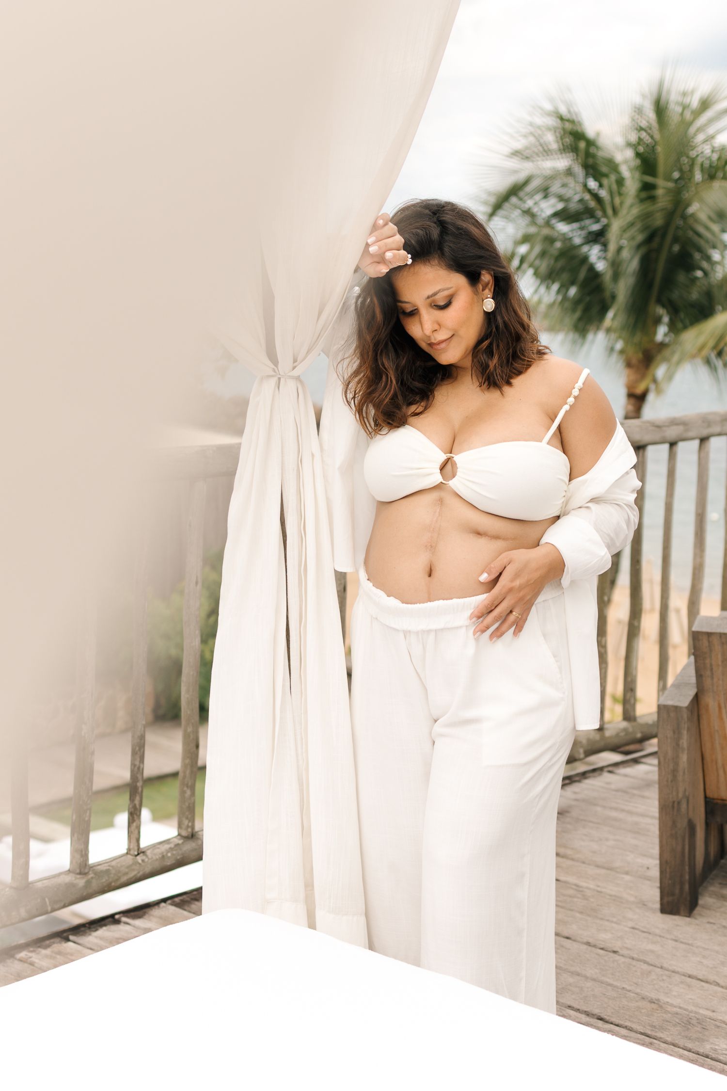 Woman in white lingerie and robe poses on wooden deck with palm trees in tropical setting at sunset.