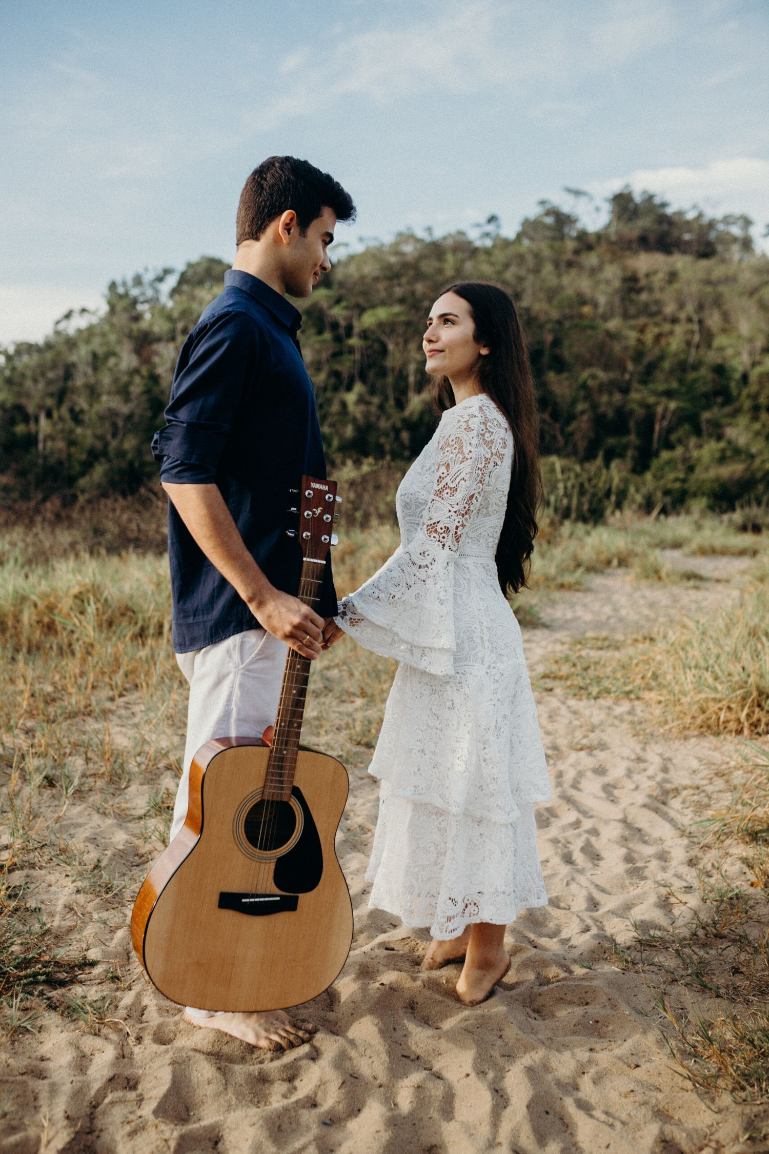 A couple poses on sandy terrain with an acoustic guitar and white lace dress.