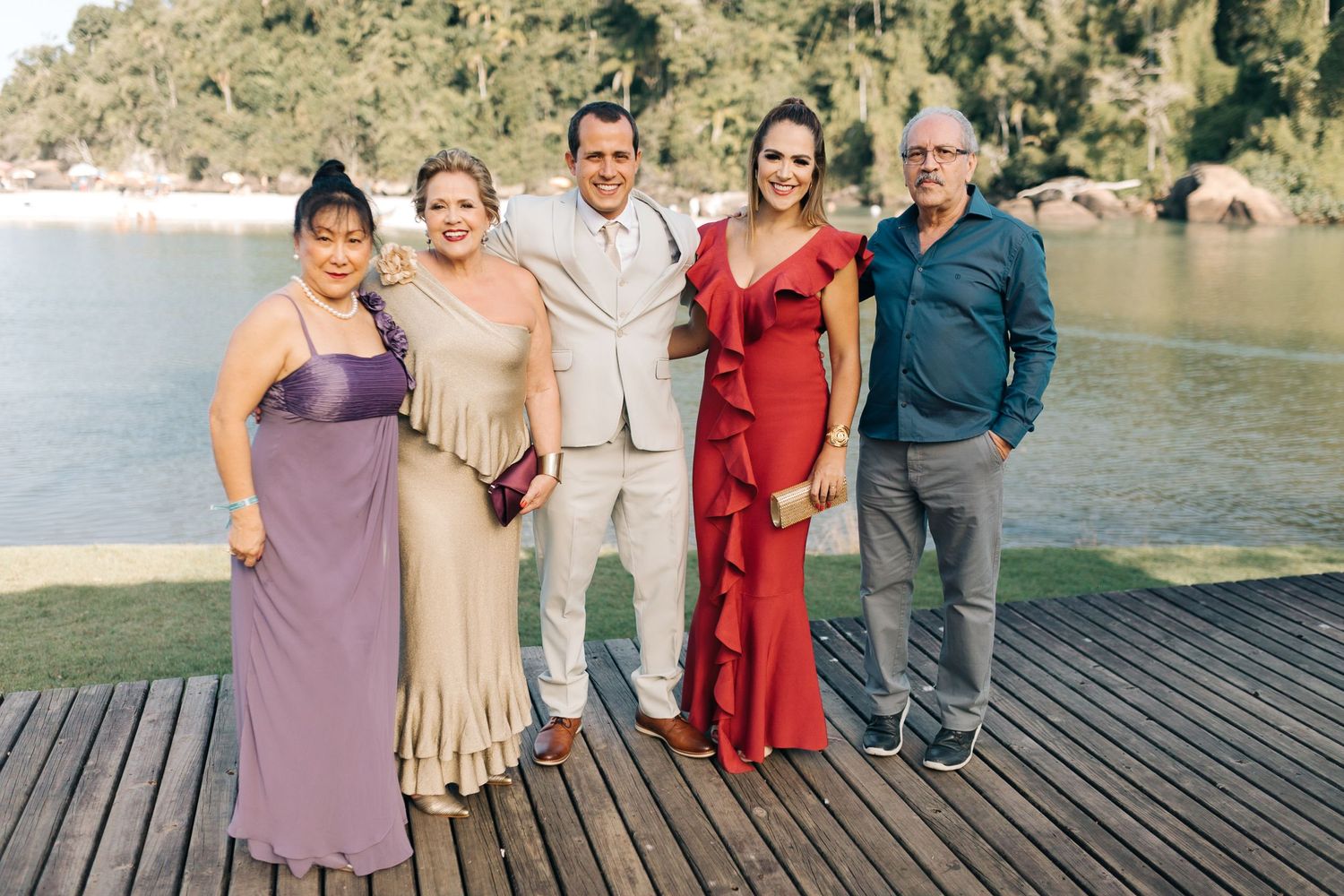 A group in formal attire poses together on a wooden deck overlooking a scenic lake.