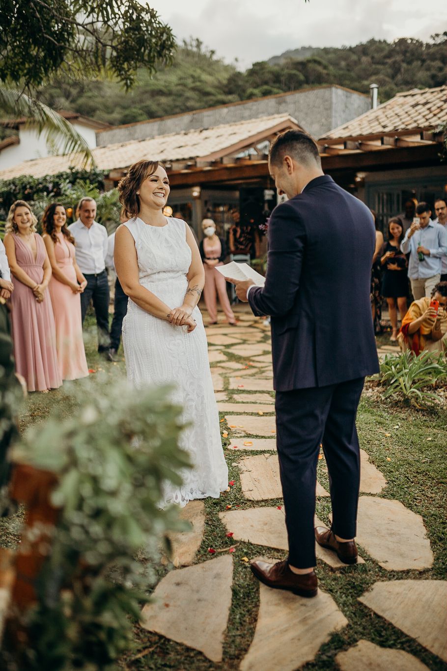 Newlyweds exchange vows on a stone pathway surrounded by lush greenery and wedding guests.