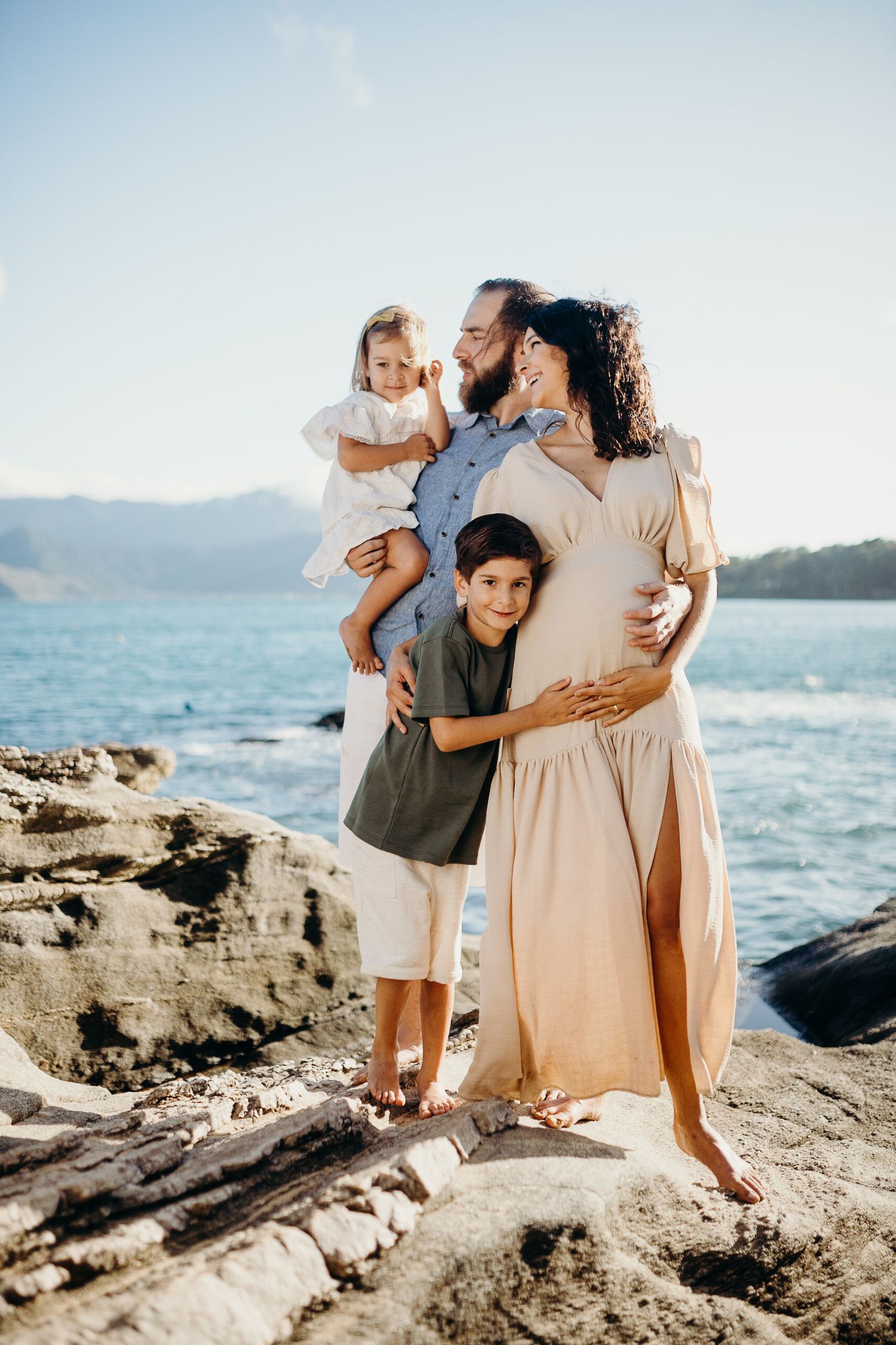 Family embraces while standing together on rocky coastal formation by ocean.