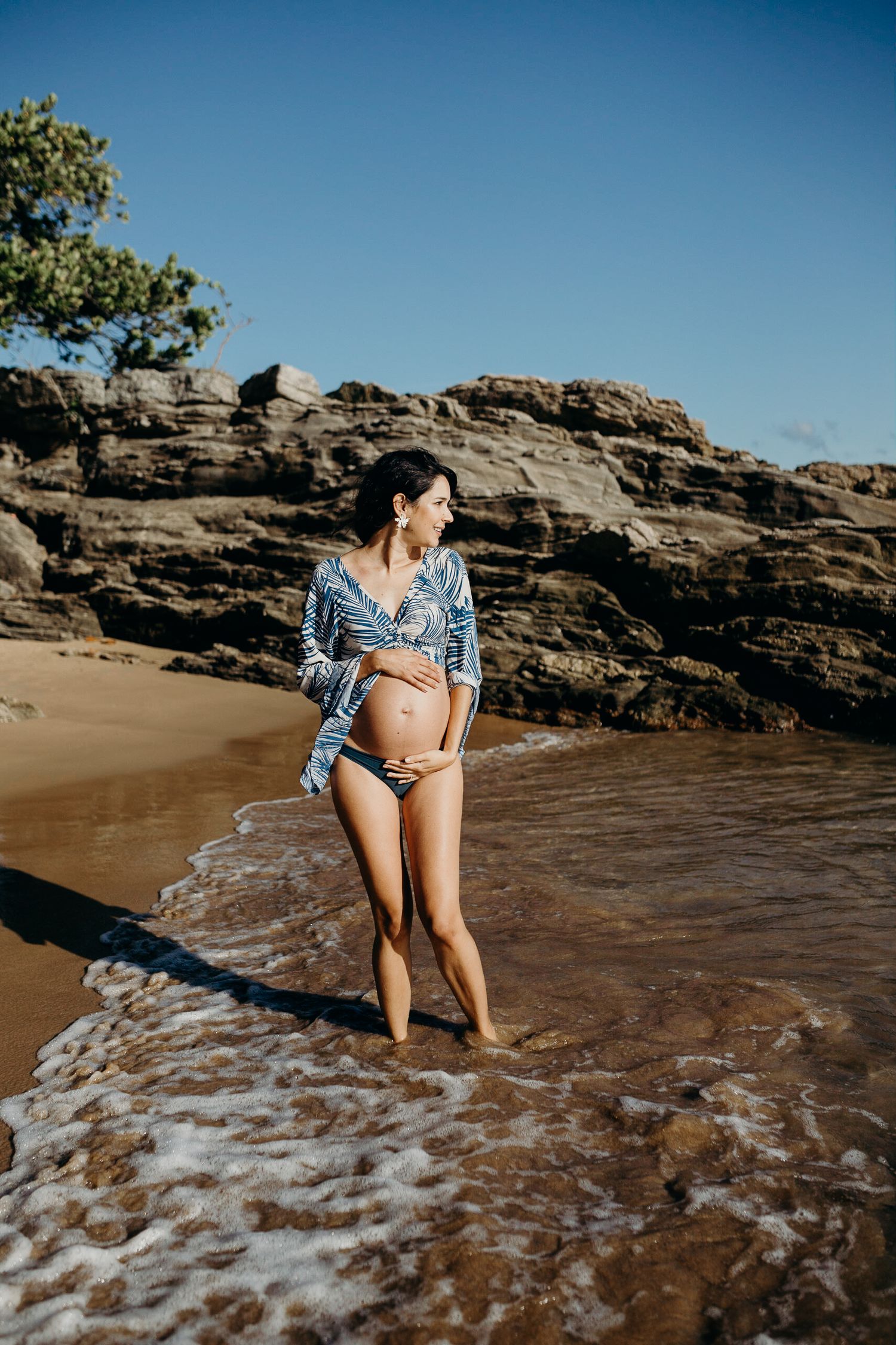 A maternity photoshoot on a rocky beach with blue waters and rocks in the background.