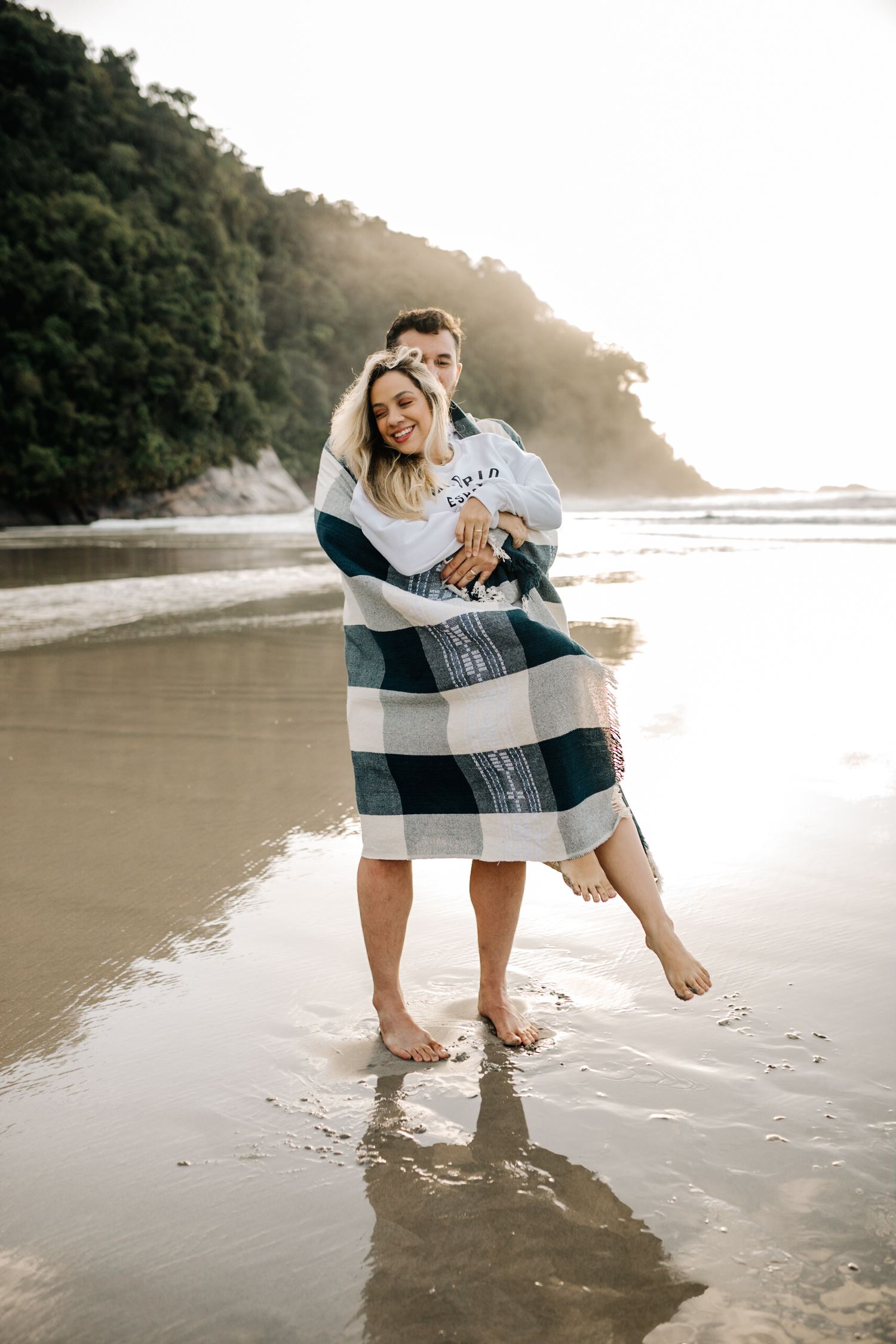 Two people share tender moments on a misty beach while wrapped in a cozy blanket.