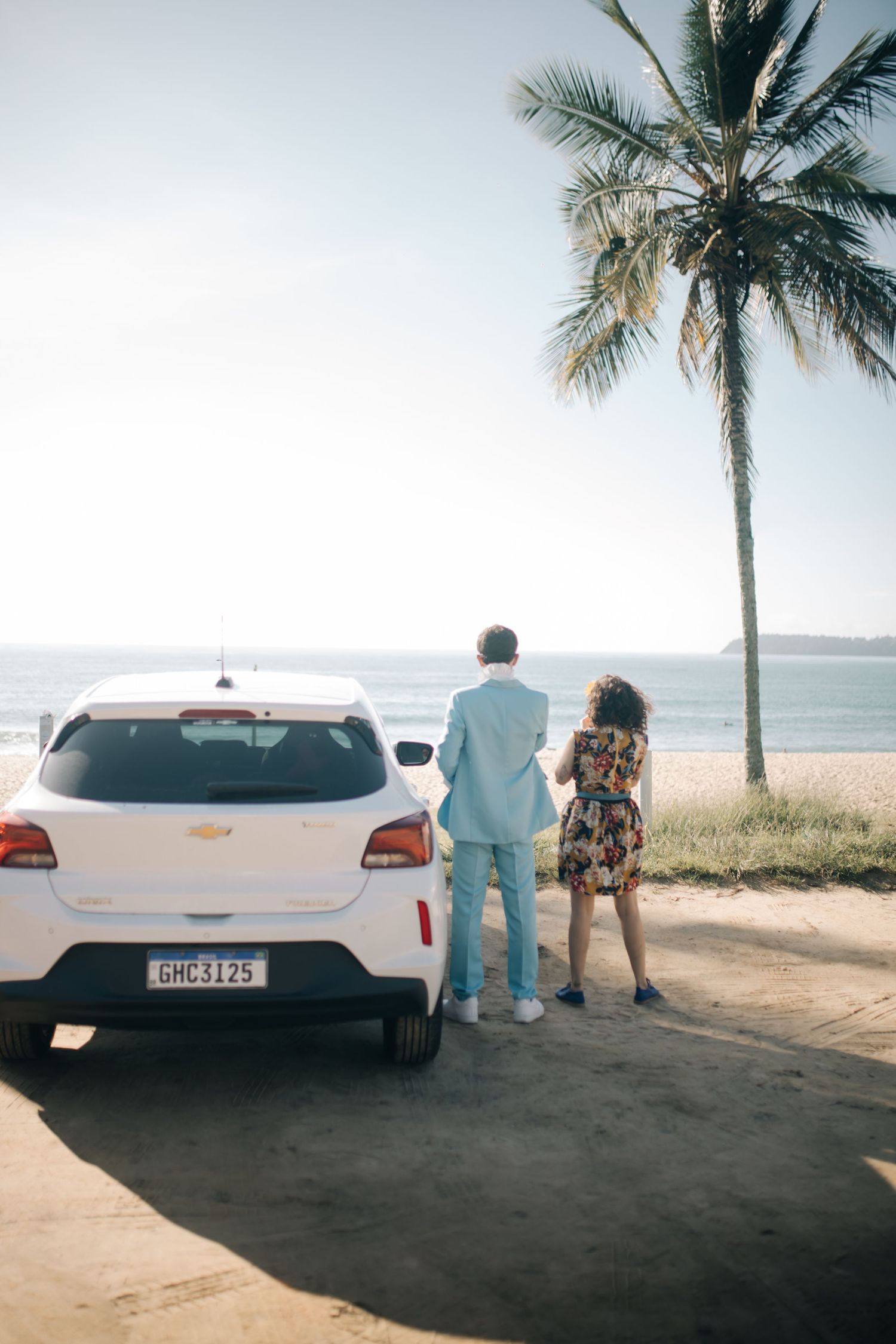 Couple standing by white car near palm tree overlooking tropical beach.
