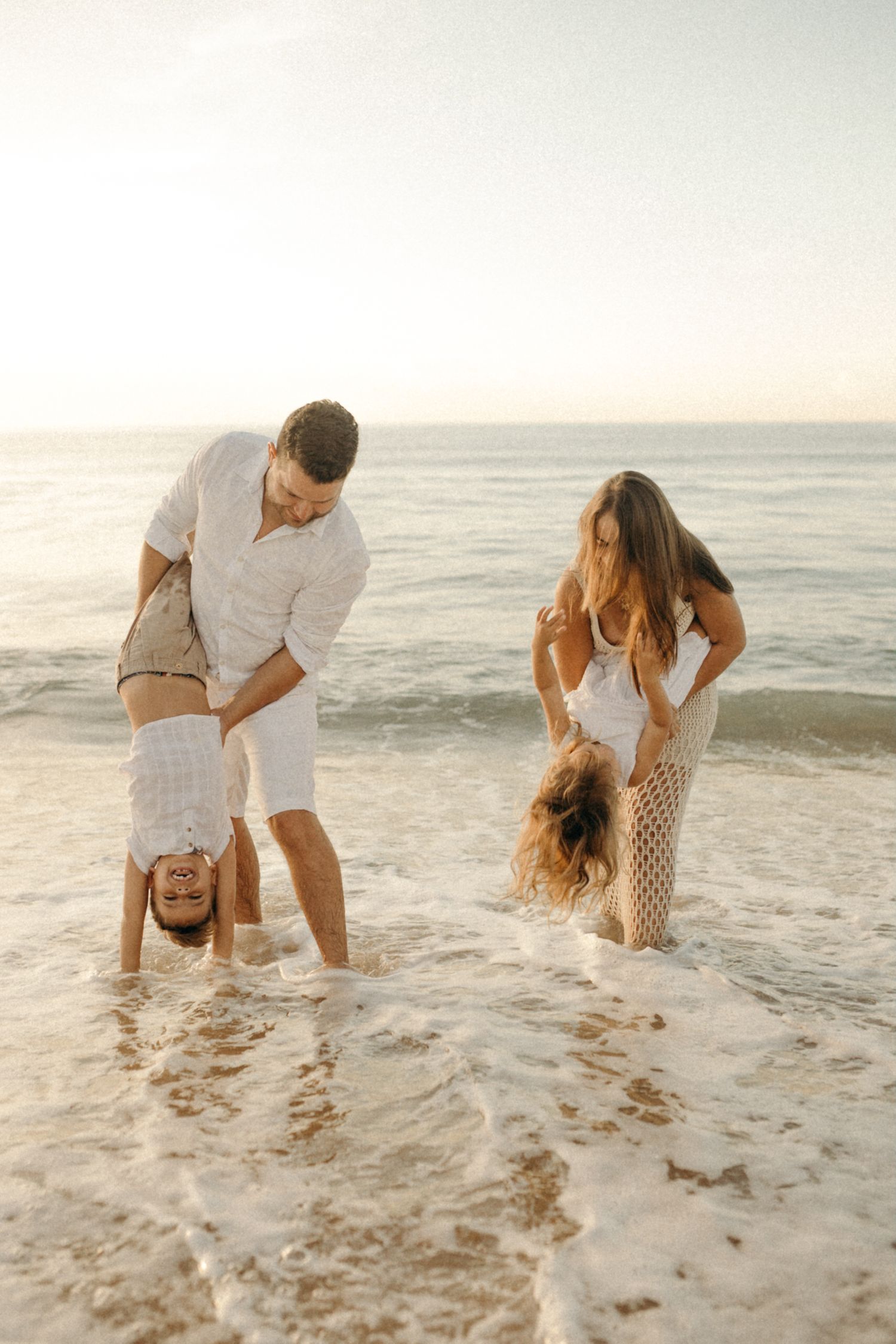 Family playing and doing handstands in shallow ocean waves at sunset.