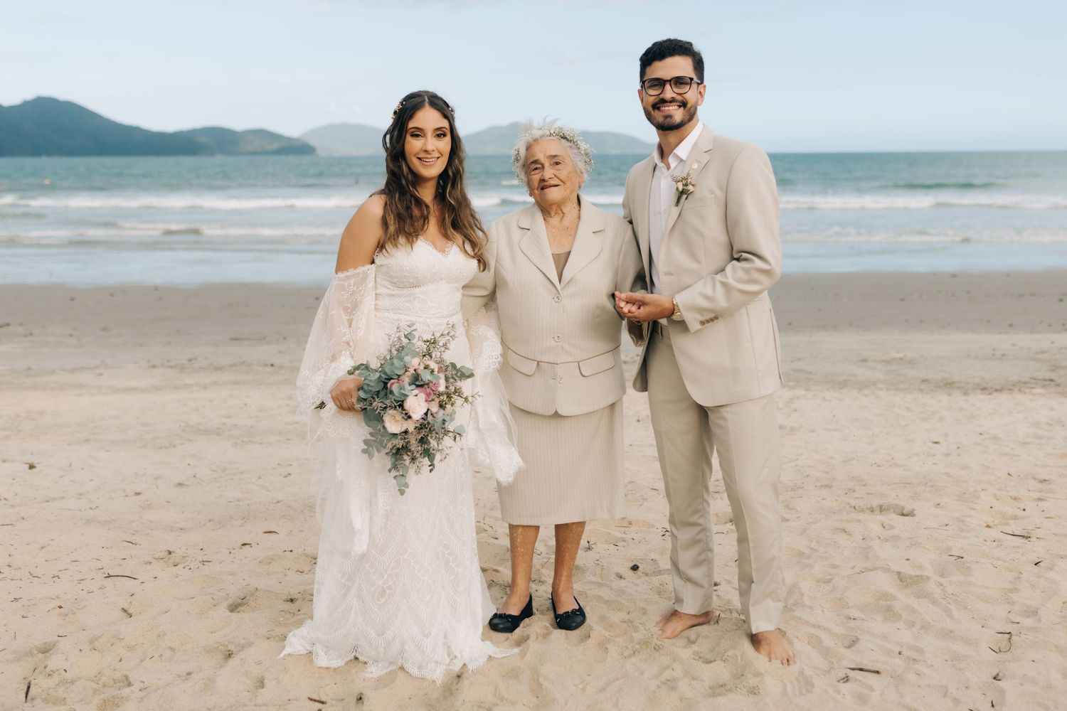 Wedding party poses for beach photo with elegant attire and floral bouquet against ocean backdrop.