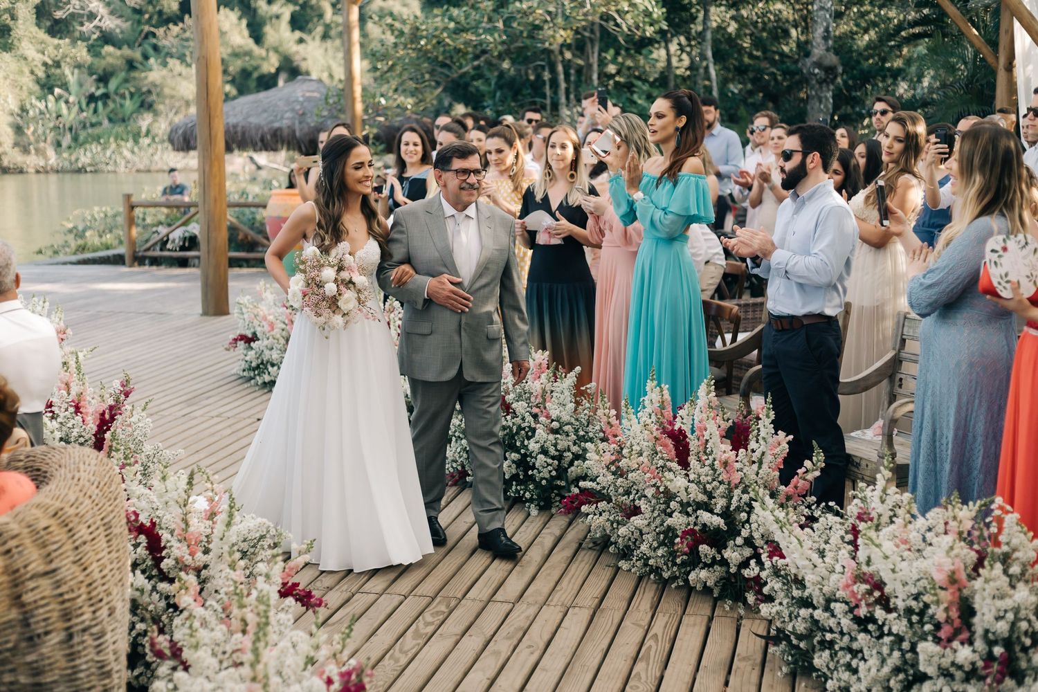 Wedding celebration on wooden deck with floral decorations and guests in pastel attire.