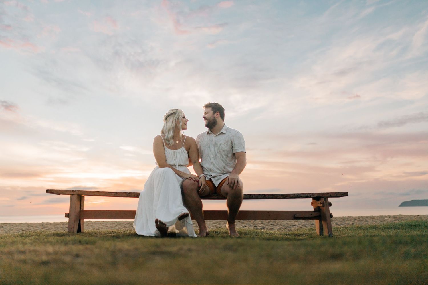 A couple sits together on a wooden bench at sunset overlooking a grassy coastal landscape.