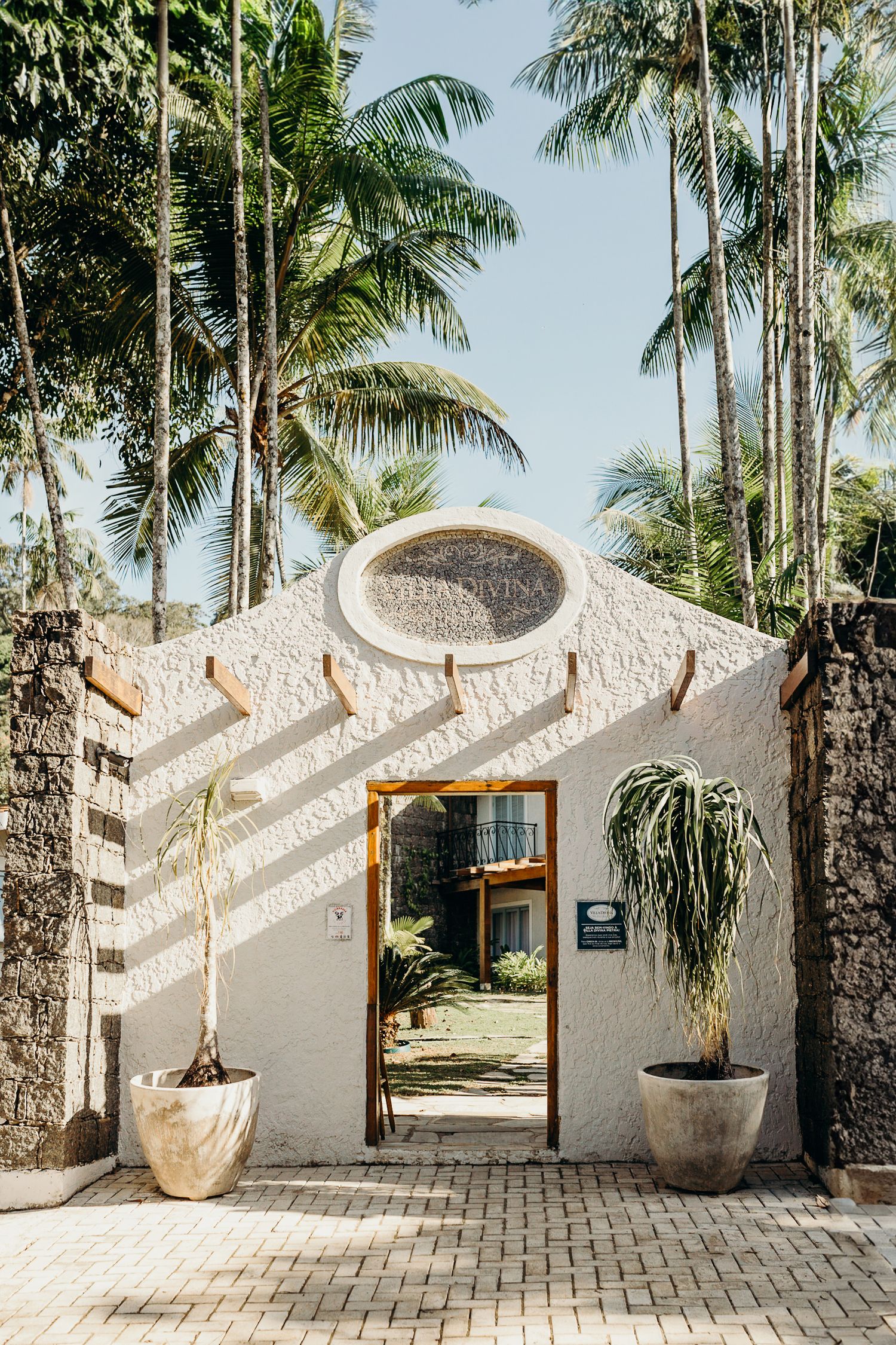 White stucco archway entrance with palm trees and potted plants in a tropical setting.