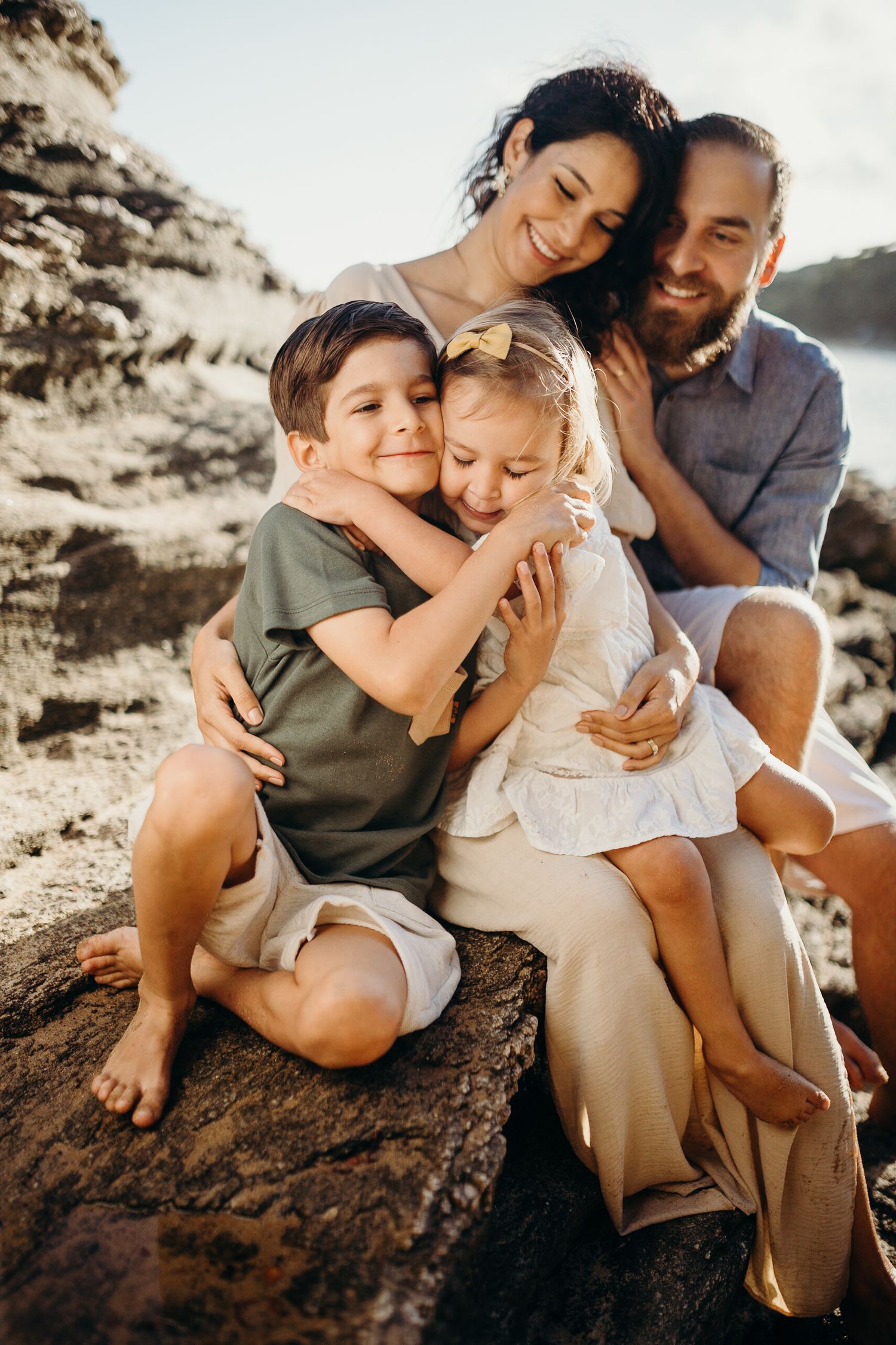 A family cuddles close together while sitting on coastal rocks during golden hour.