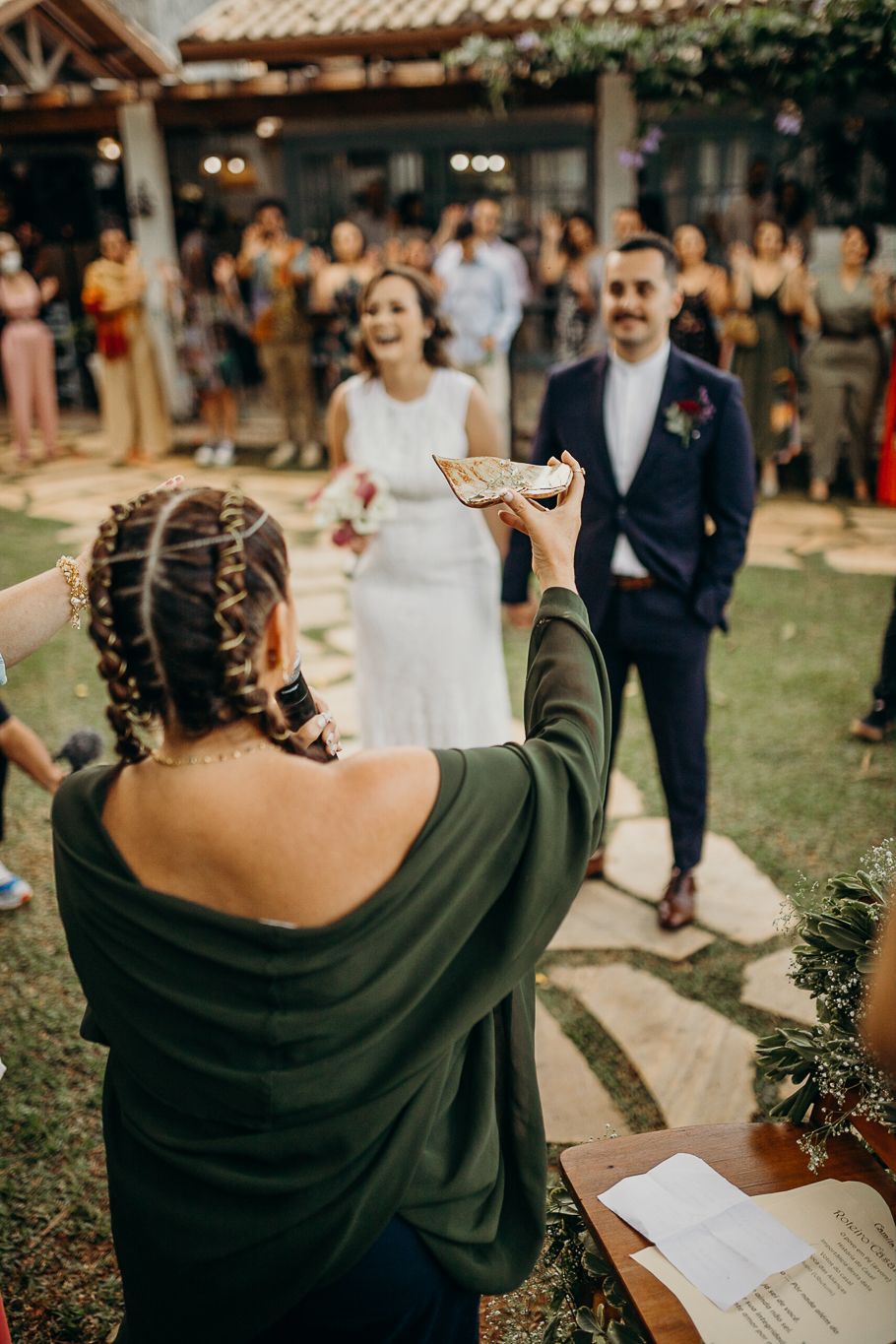 An outdoor wedding ceremony with an officiant addressing a couple while holding a book at a garden venue.