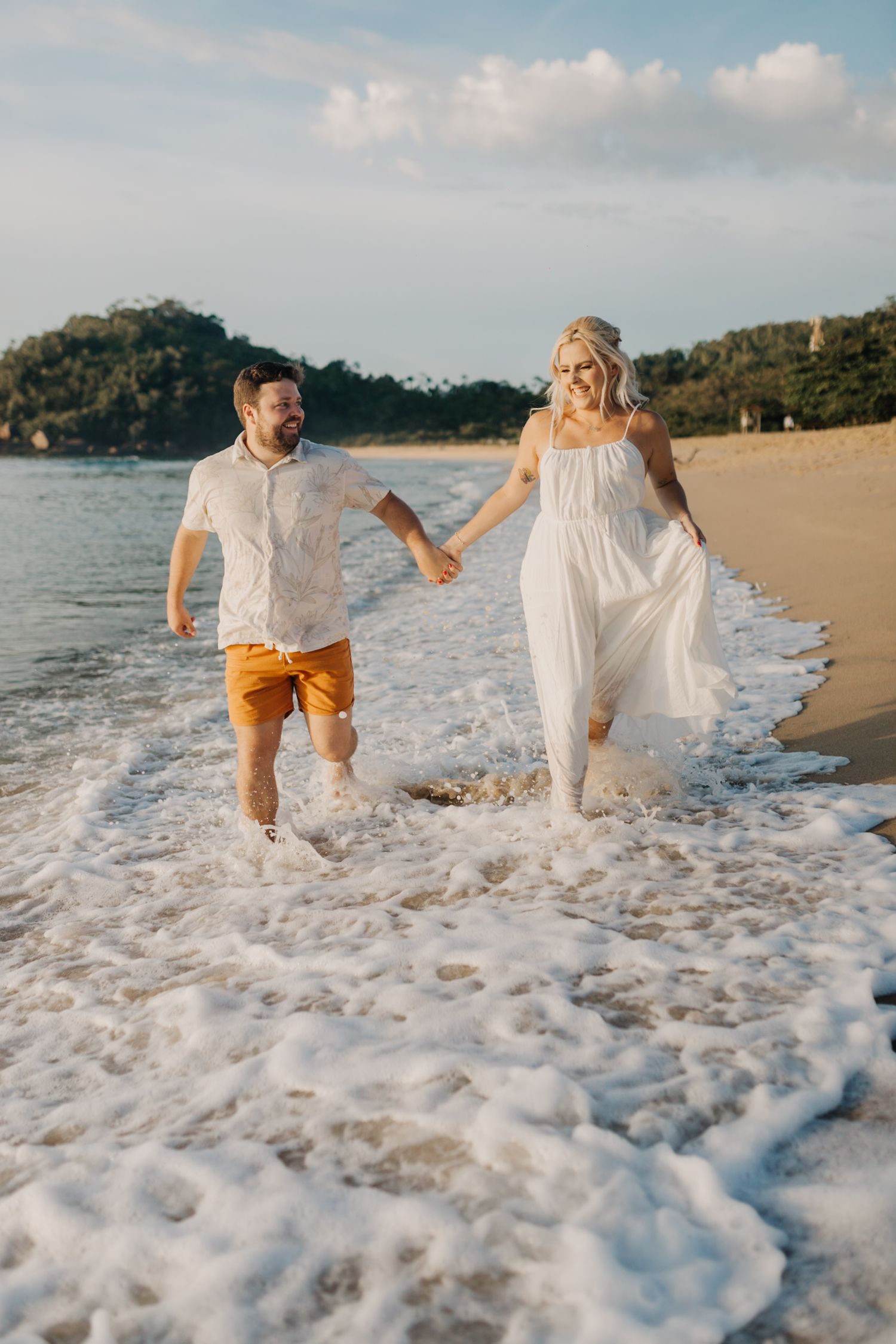 Couple splashing through ocean waves in formal beach attire during sunset photoshoot.