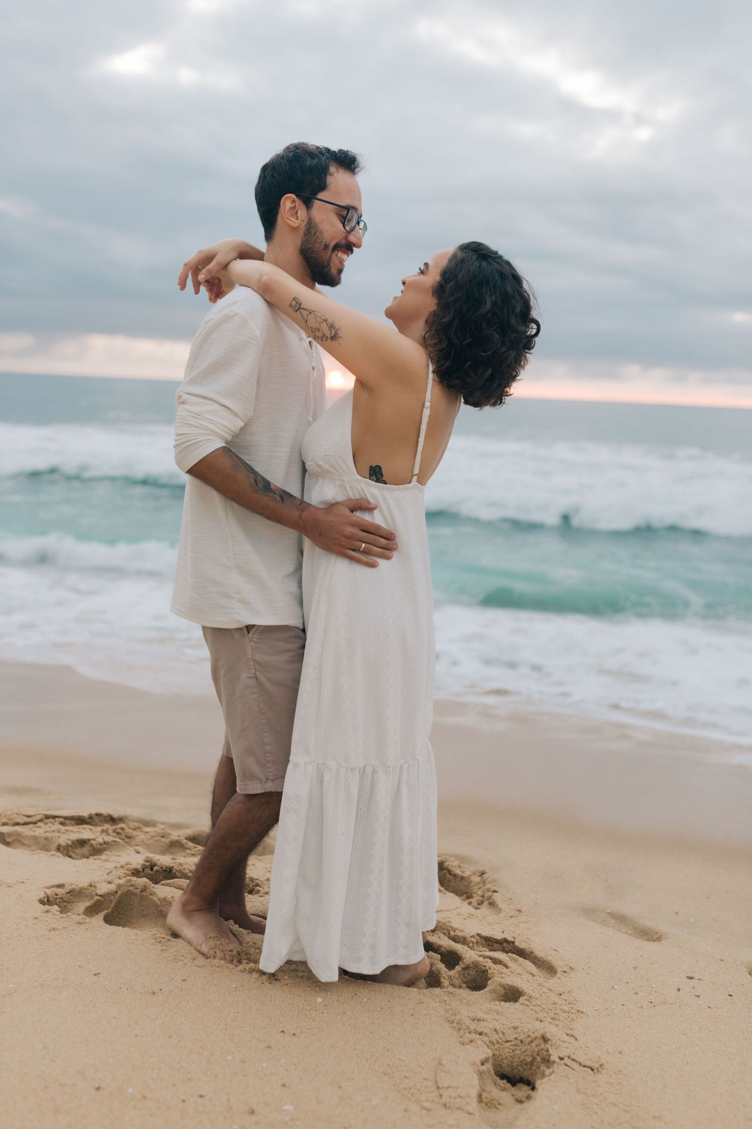 A couple embraces on the sandy beach with crashing waves and cloudy skies in the background.