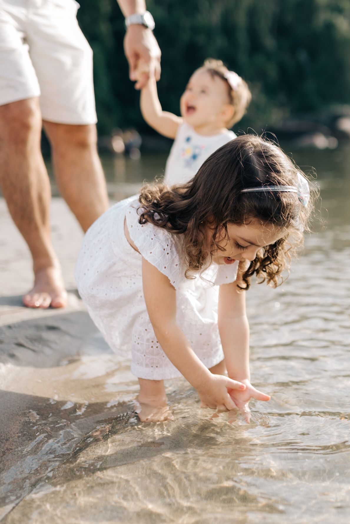A child in a white dress explores the shallow water while an adult watches nearby.