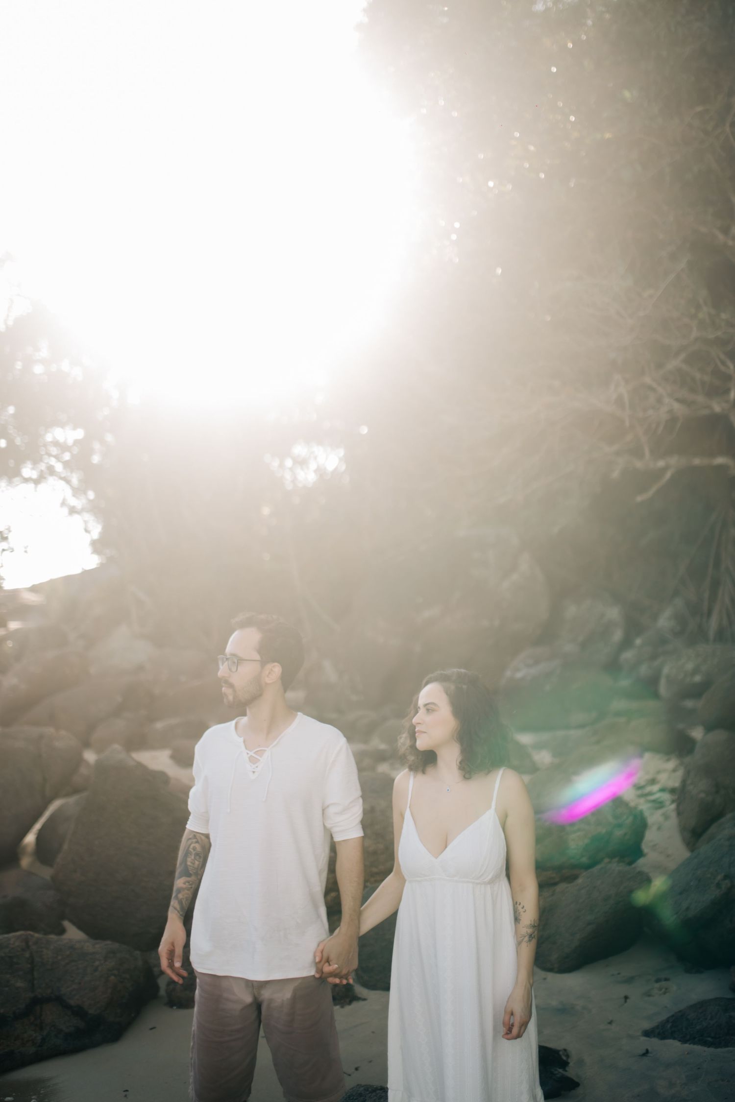 Couple in white outfits standing together on rocky beach during golden hour sunset.