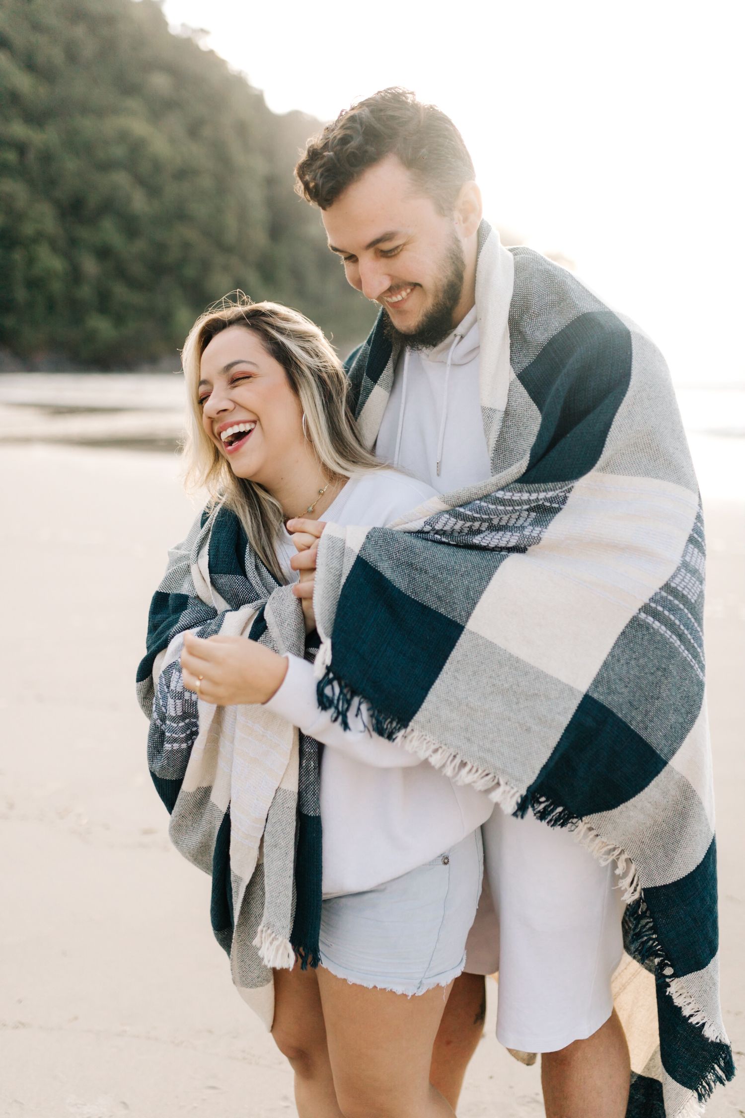 A happy couple laughs together while sharing a cozy striped blanket on the beach.