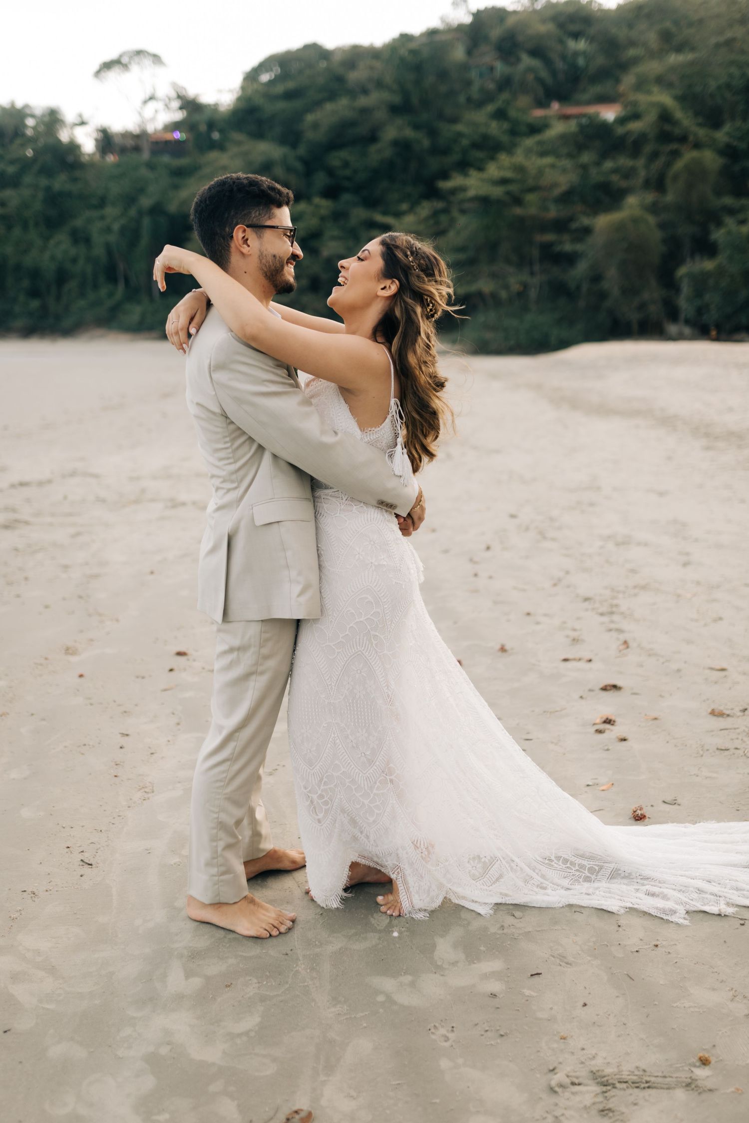 A couple embraces on a sandy beach, with the bride in white lace and groom in beige.