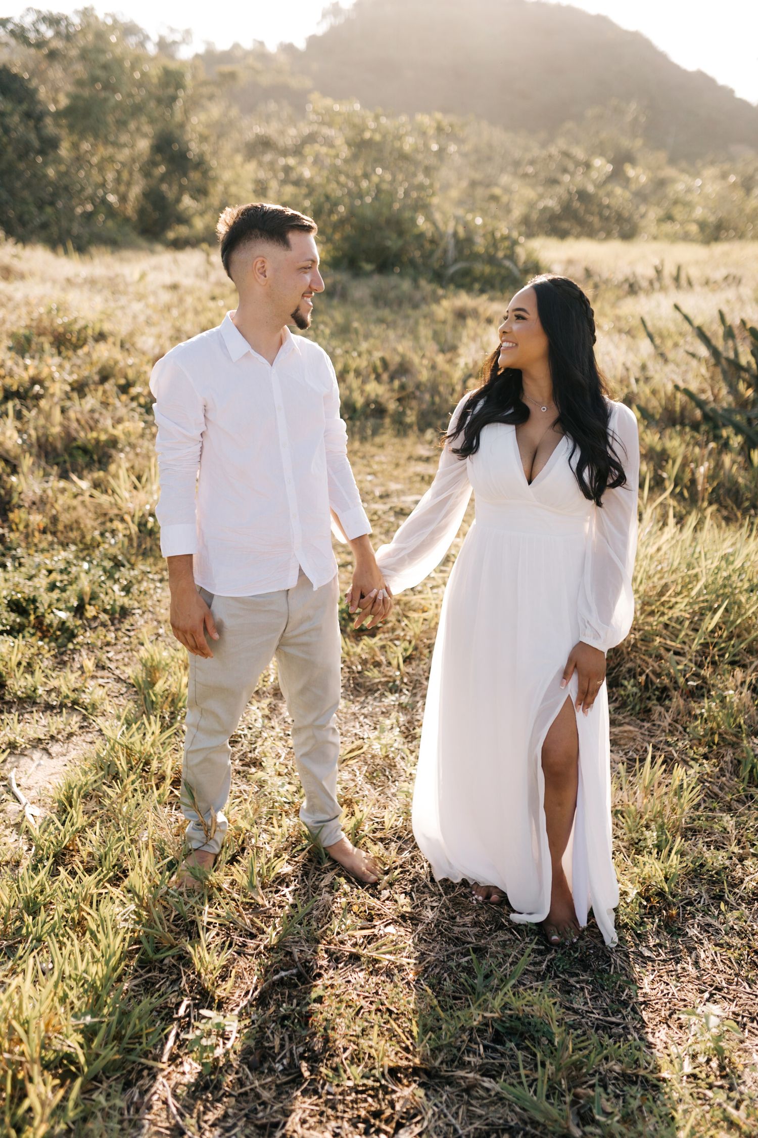 A romantic couple shares a joyful moment in a sunlit meadow wearing coordinated white and beige outfits.