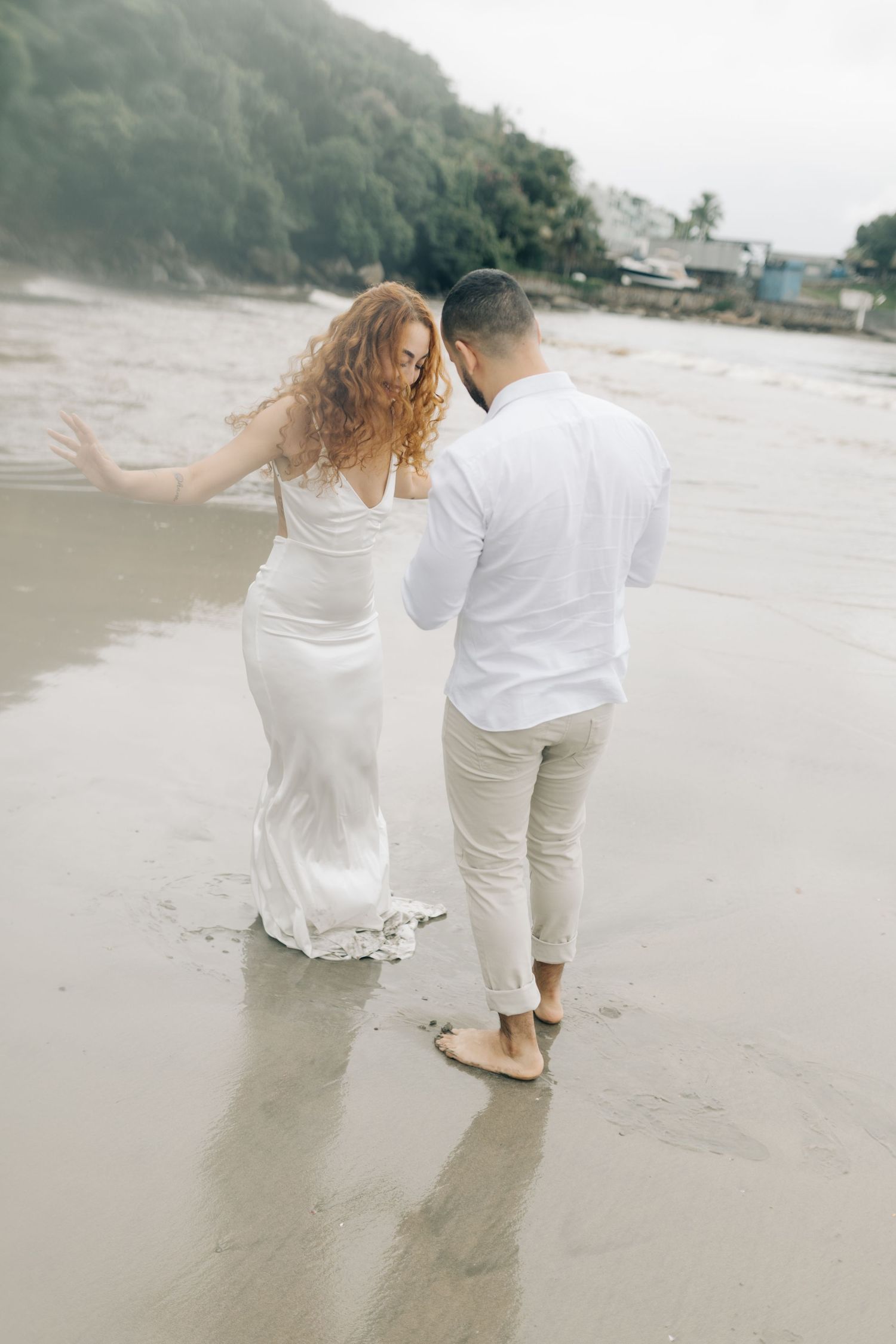 A couple in white attire shares a joyful moment walking along a misty beach shoreline.