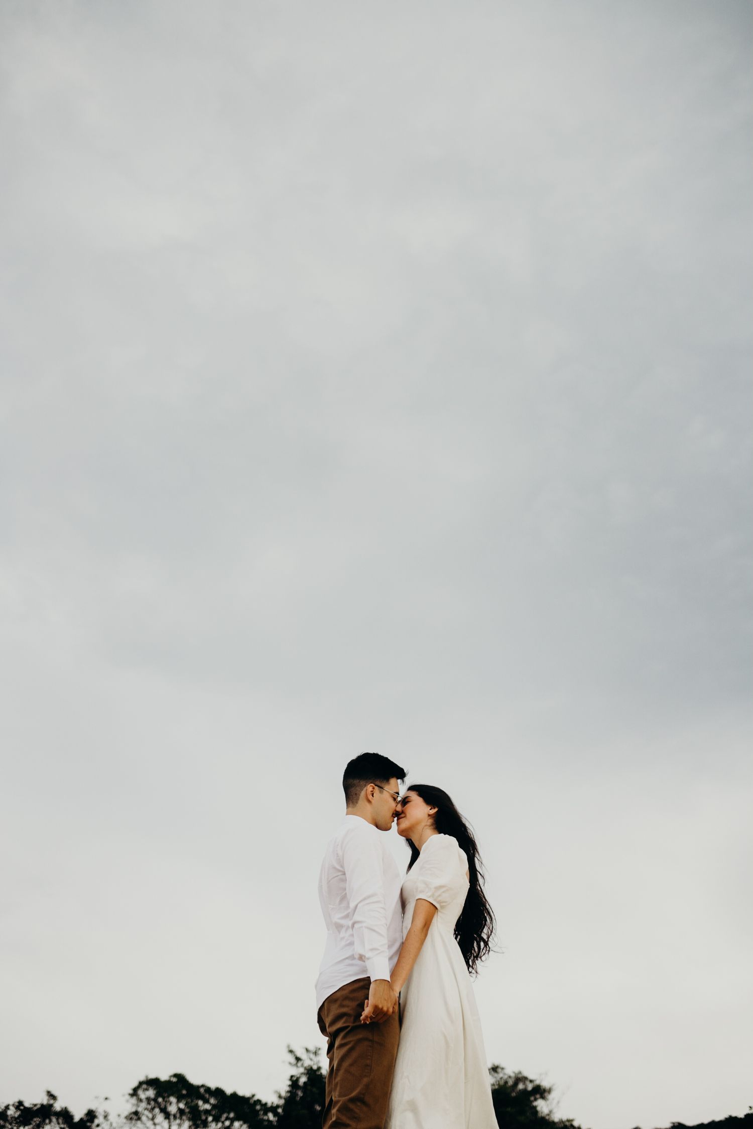 A couple embraces in silhouette against a cloudy sky with trees in the background.
