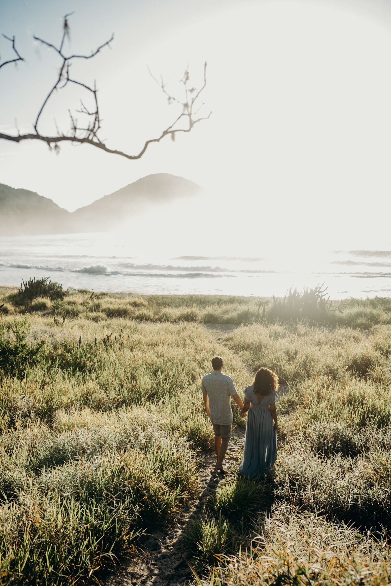 Couple walking through coastal grassland with misty ocean view in background.