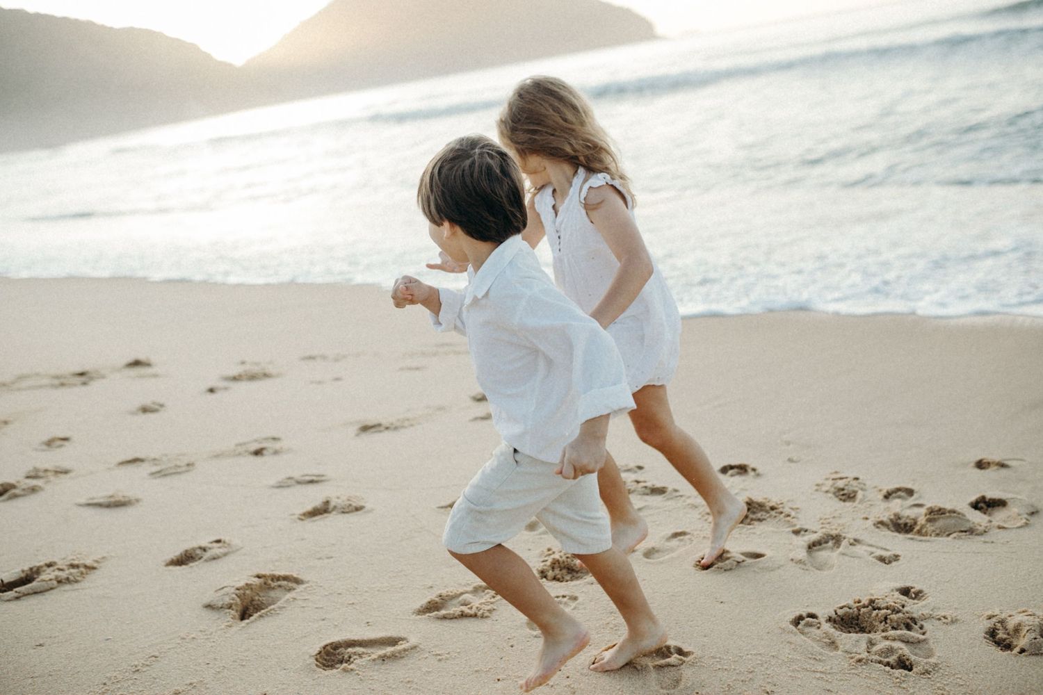 Two people in white outfits running together on a beach with mountains in the background.