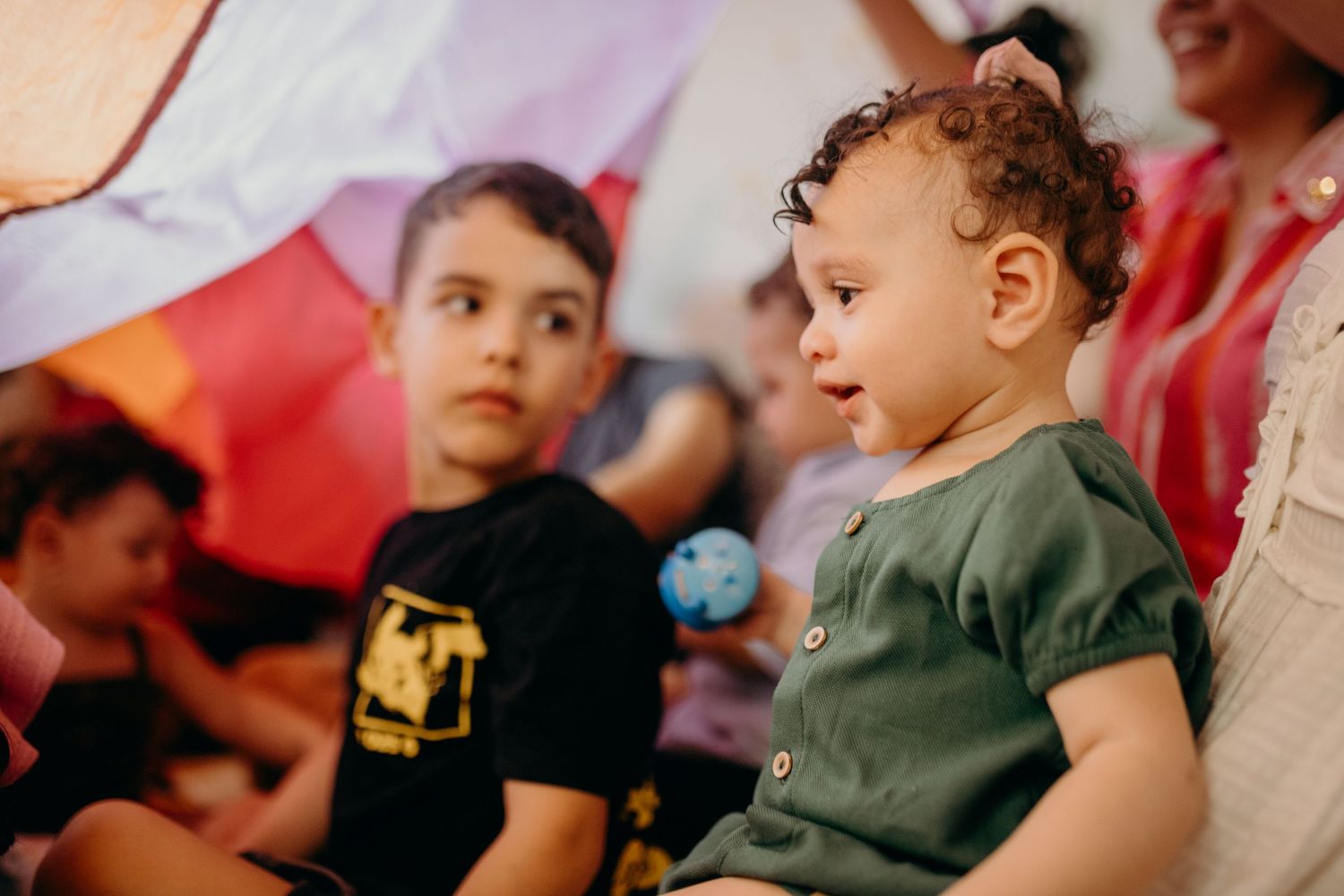 Children watch attentively during an indoor group activity with colorful fabric overhead.